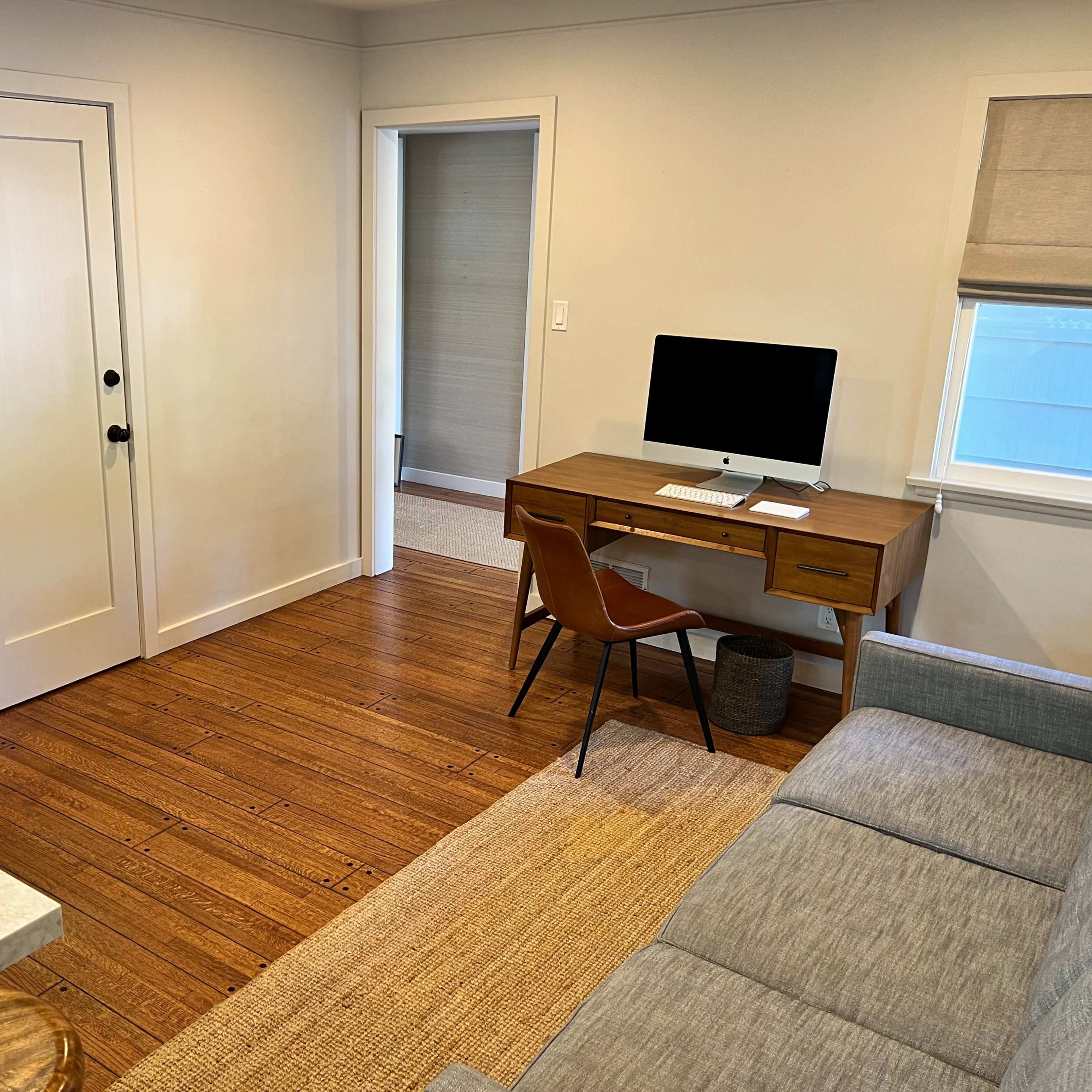 A modern living space features a desk with an iMac, a chair, and a couch, along with hardwood flooring and minimal decor.