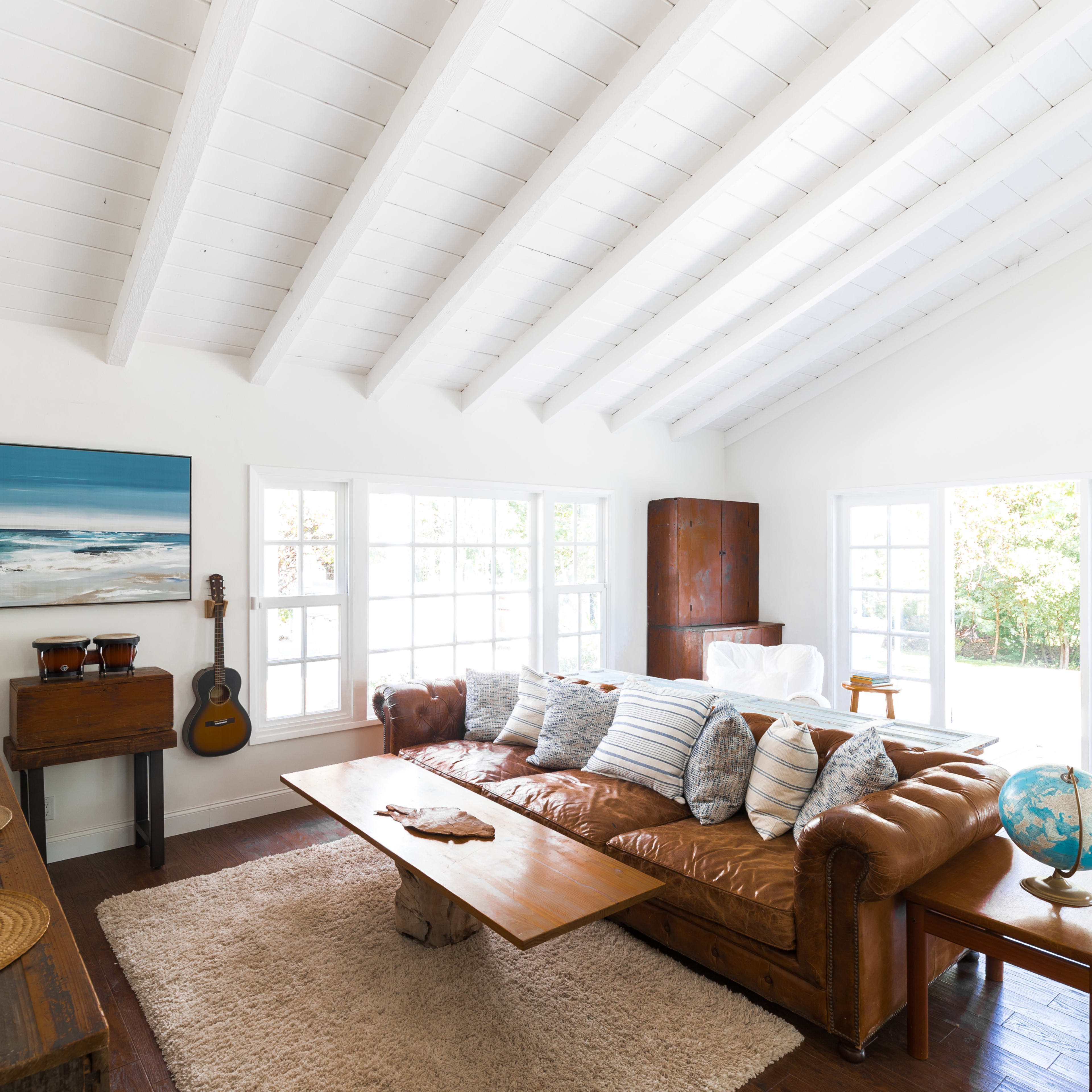 The living room features a brown leather sofa, a wooden coffee table, and large windows that allow natural light to fill the space.