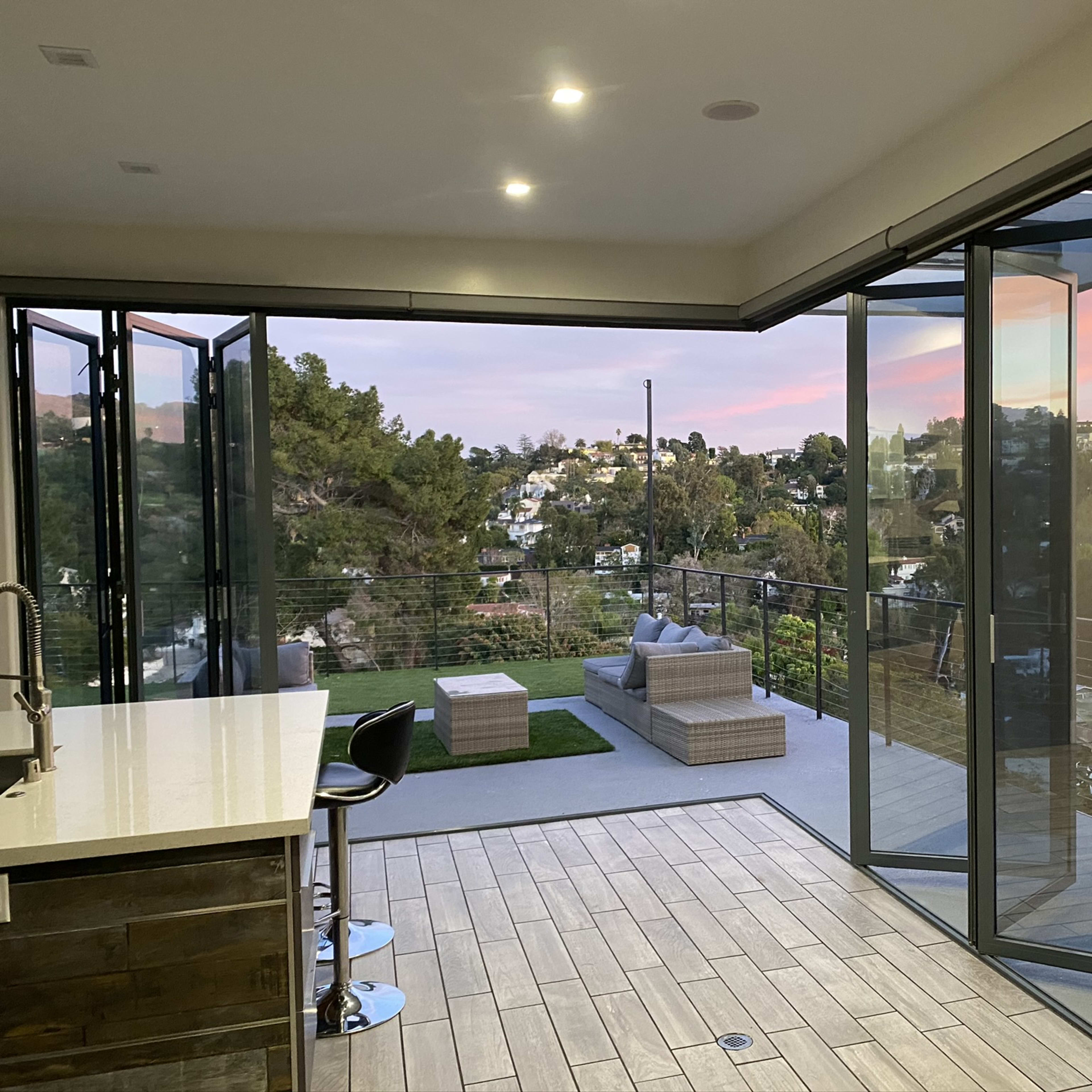 A modern indoor kitchen area with large glass doors opening to a scenic outdoor patio overlooking hills and houses at sunset.