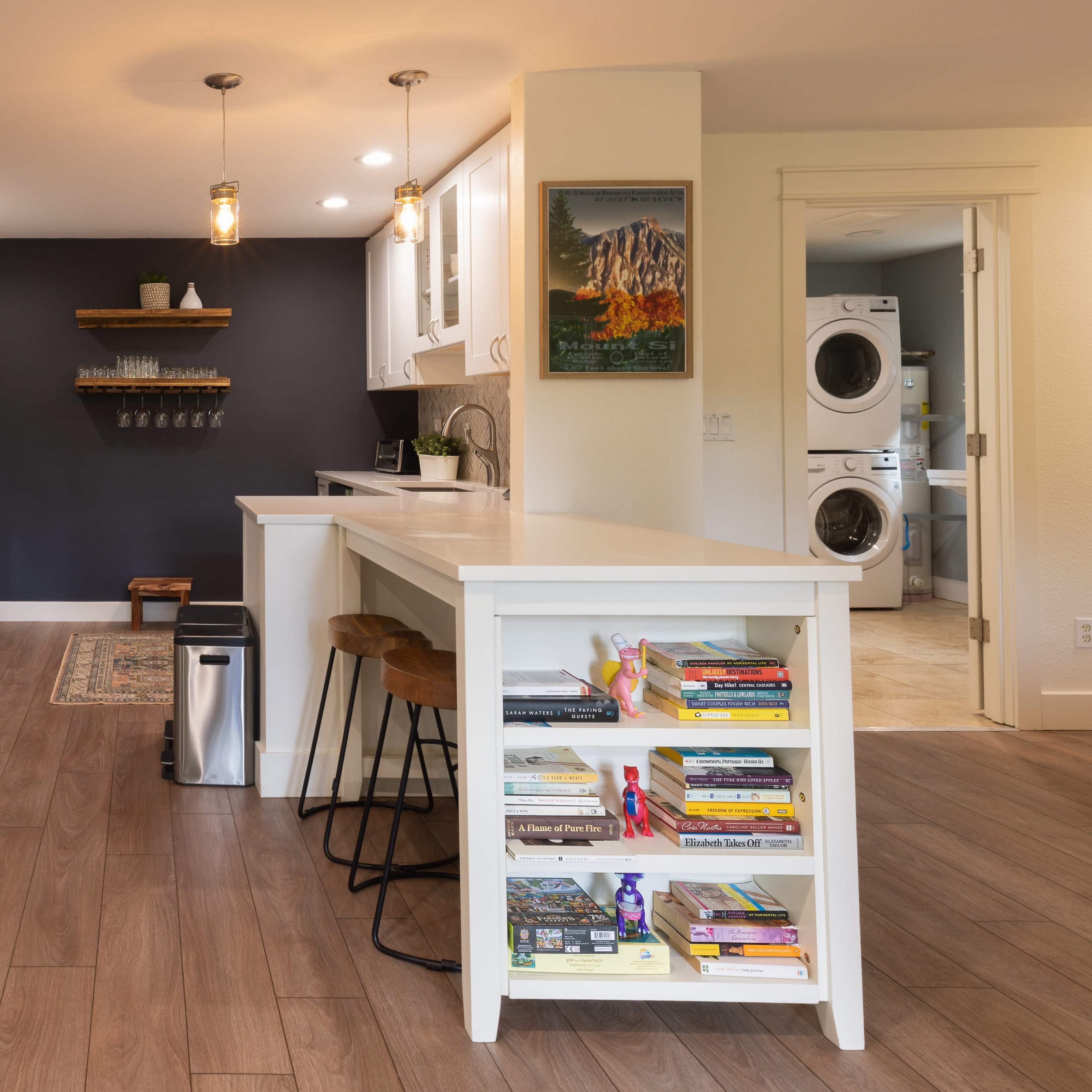 A modern kitchen features a central island with bookshelves, stainless steel appliances, and a laundry area to the side.