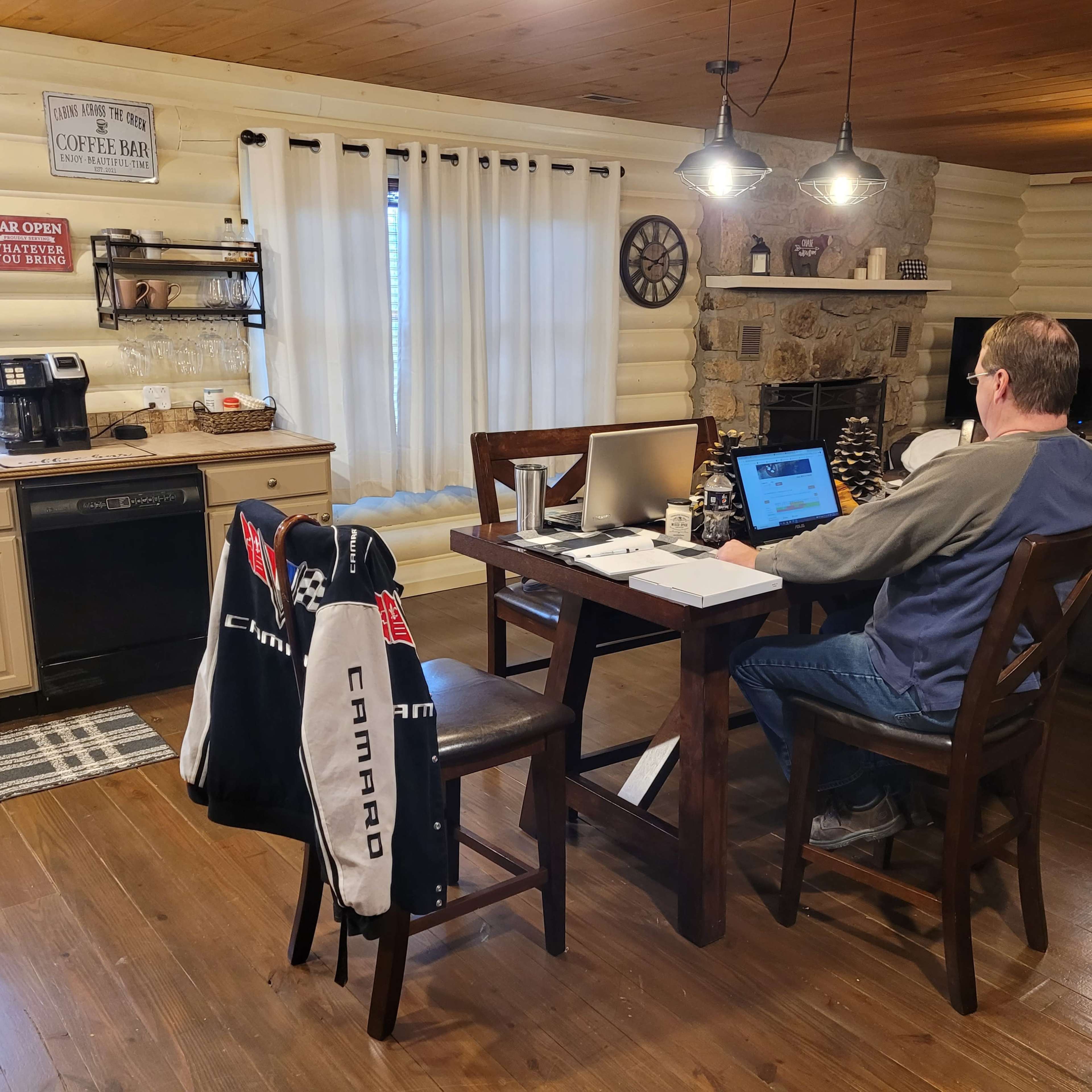 A person is working at a wooden table in a cozy kitchen with log cabin walls, a coffee maker, and a stone fireplace.