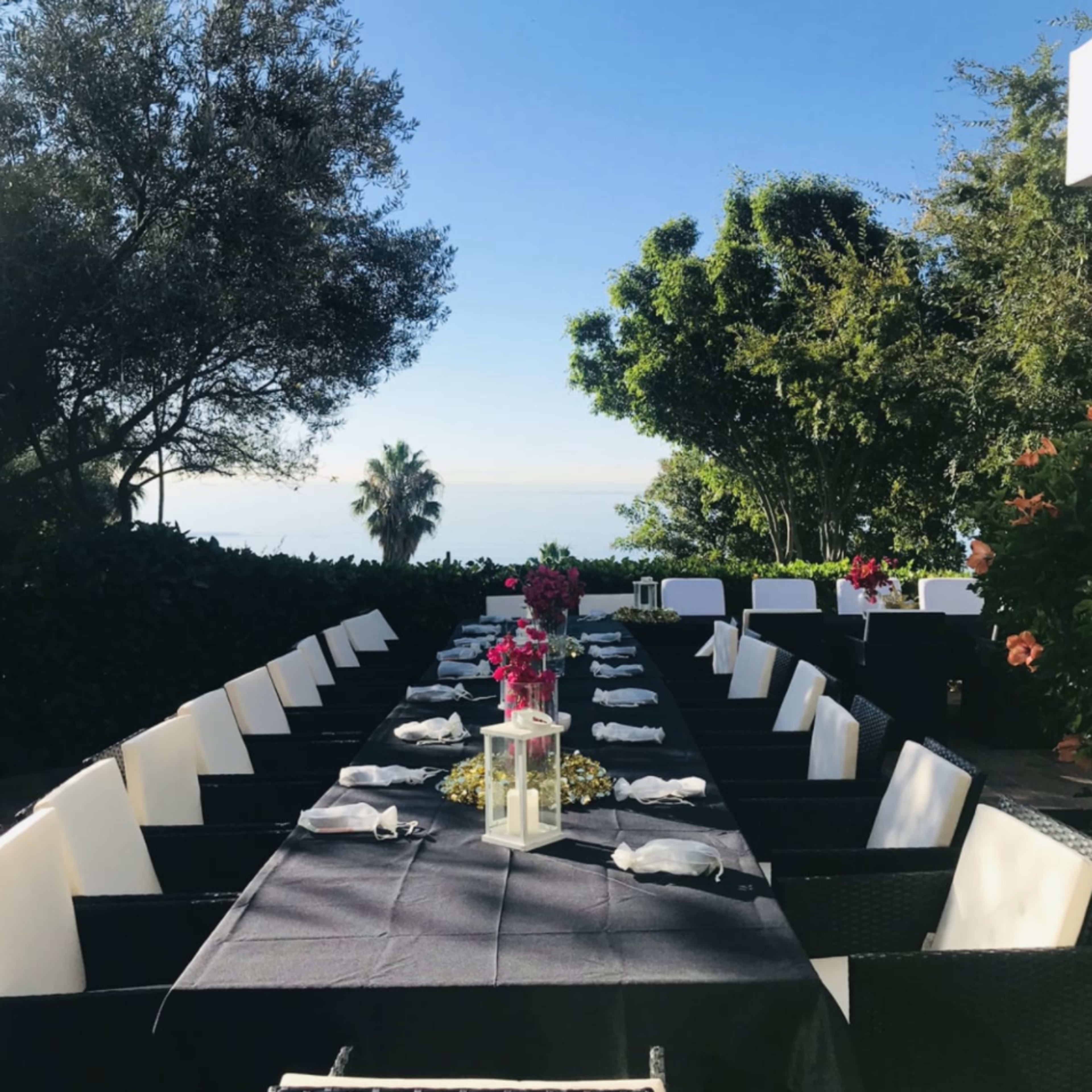 A long outdoor dining table is set with white napkins and a centerpiece, surrounded by greenery and overlooking the ocean.