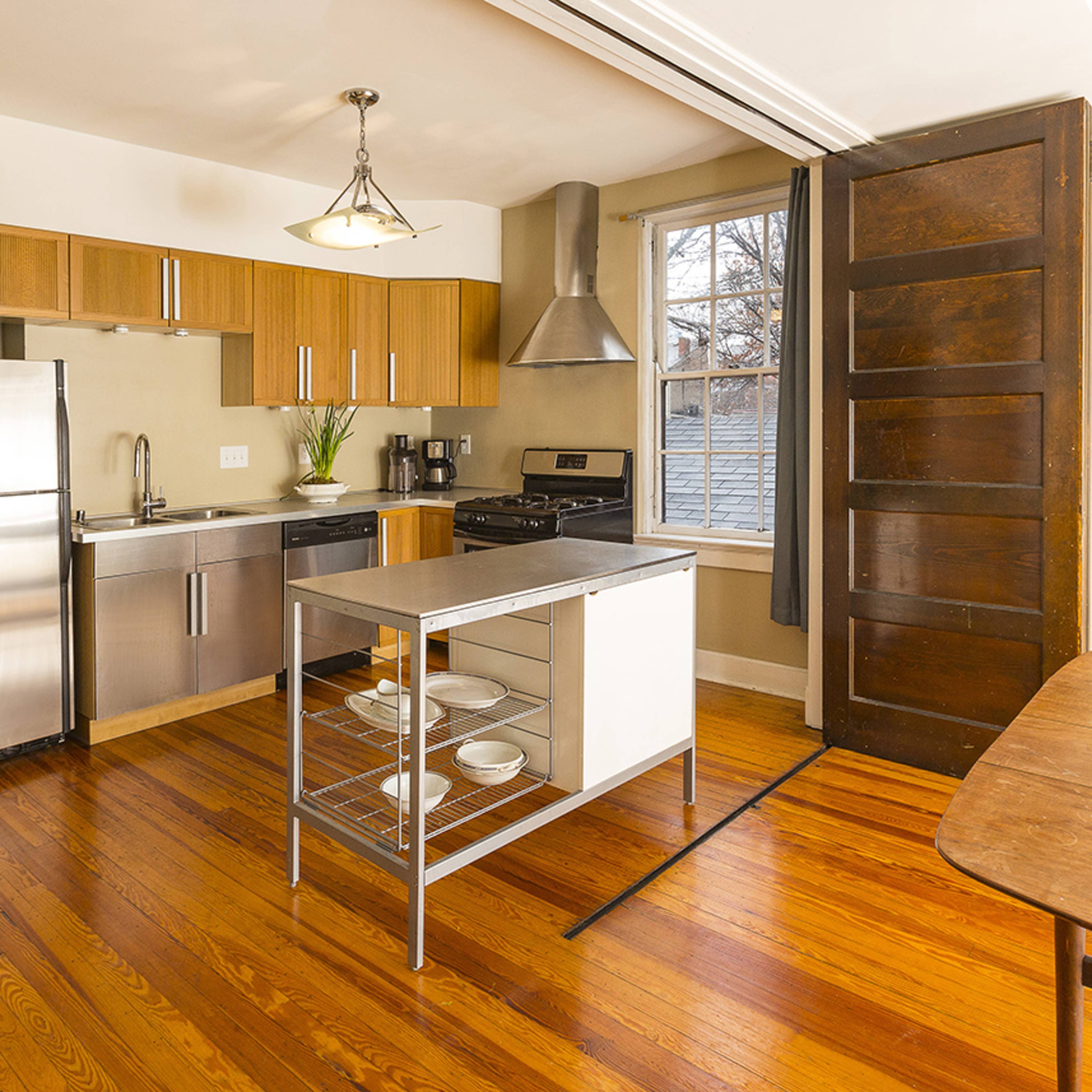 A modern kitchen featuring stainless steel appliances, wooden cabinets, and a table, with hardwood floors and a large window.
