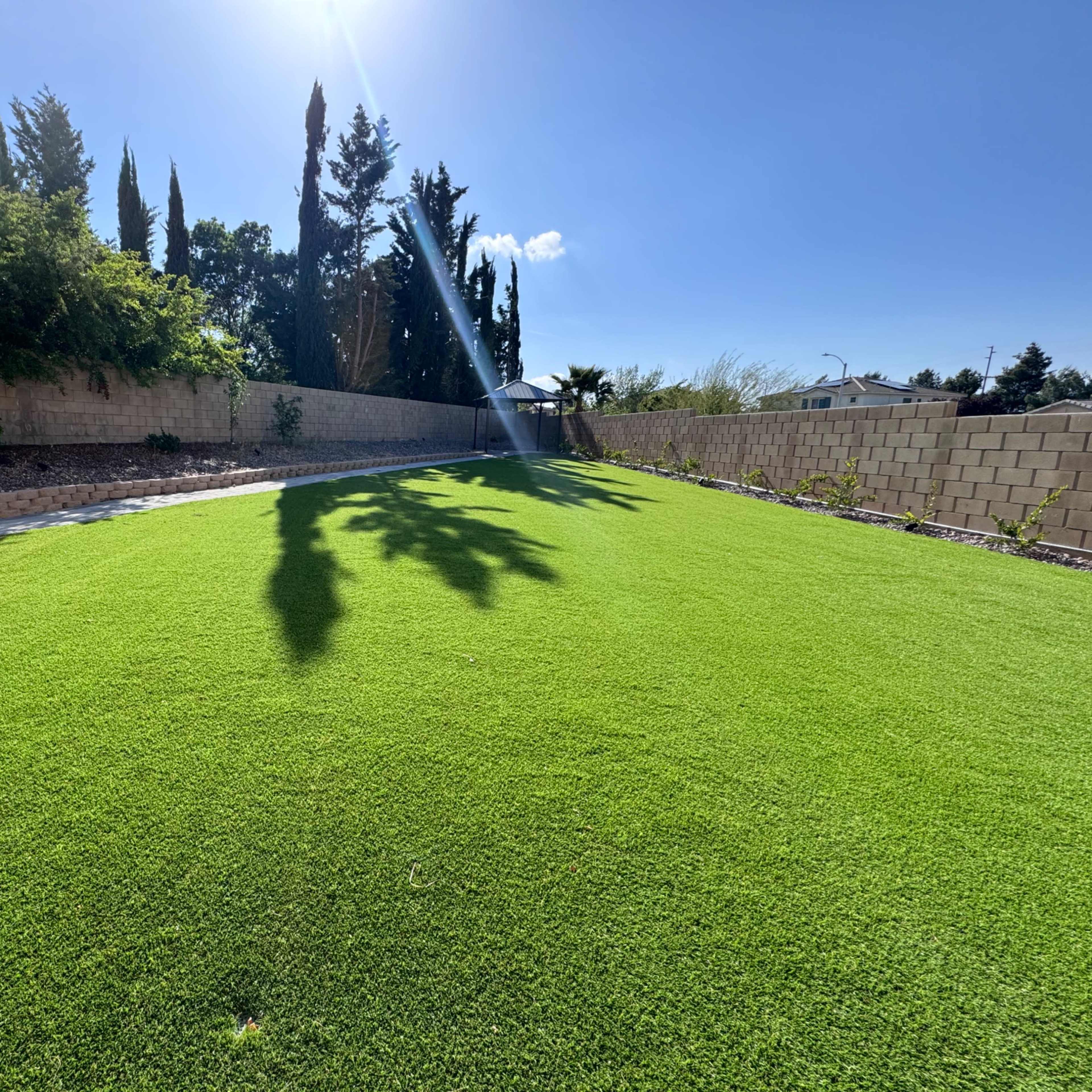 A green artificial lawn is surrounded by a low stone wall and various trees under a clear blue sky.