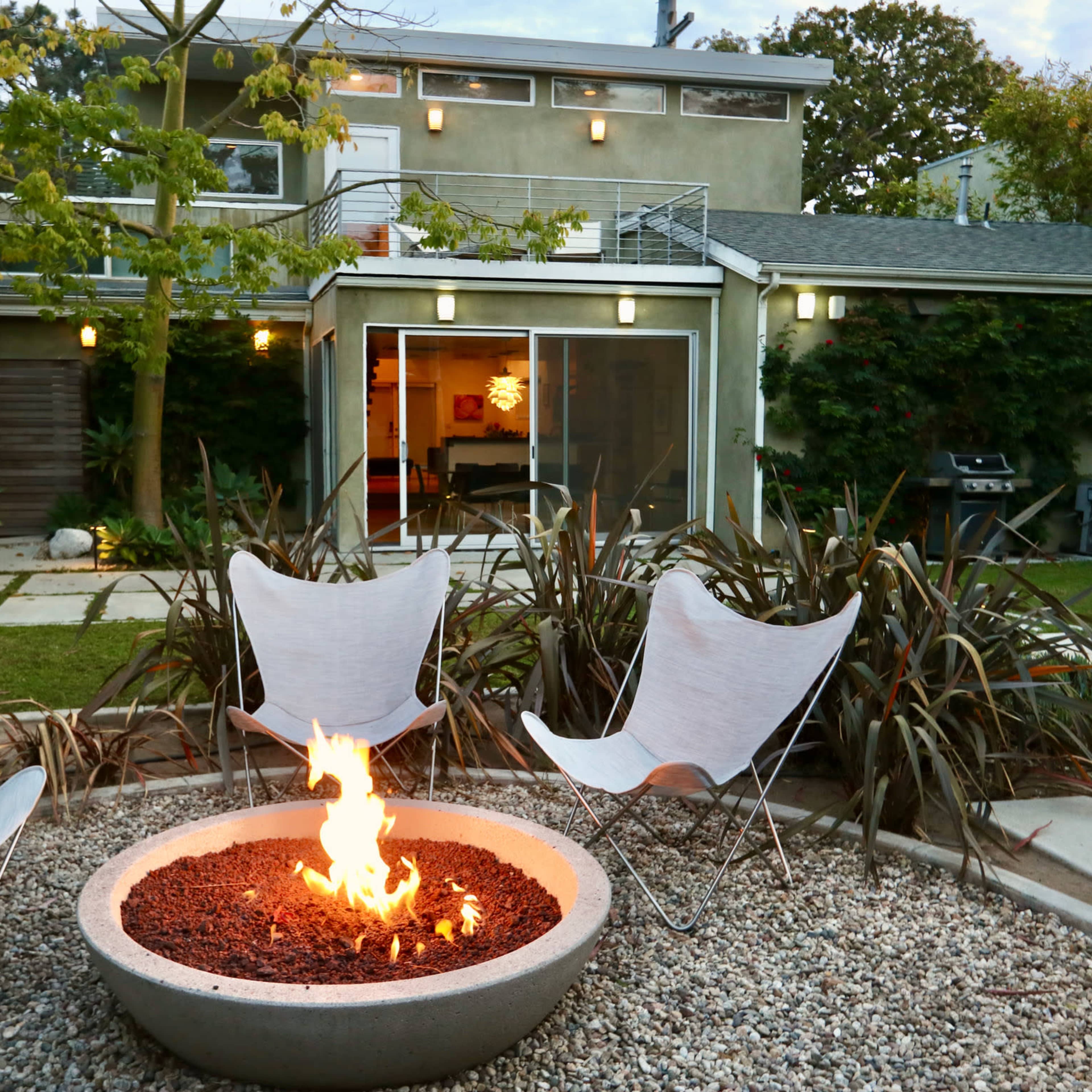 A modern backyard features a circular fire pit surrounded by four butterfly chairs, with a house and lush greenery in the background.