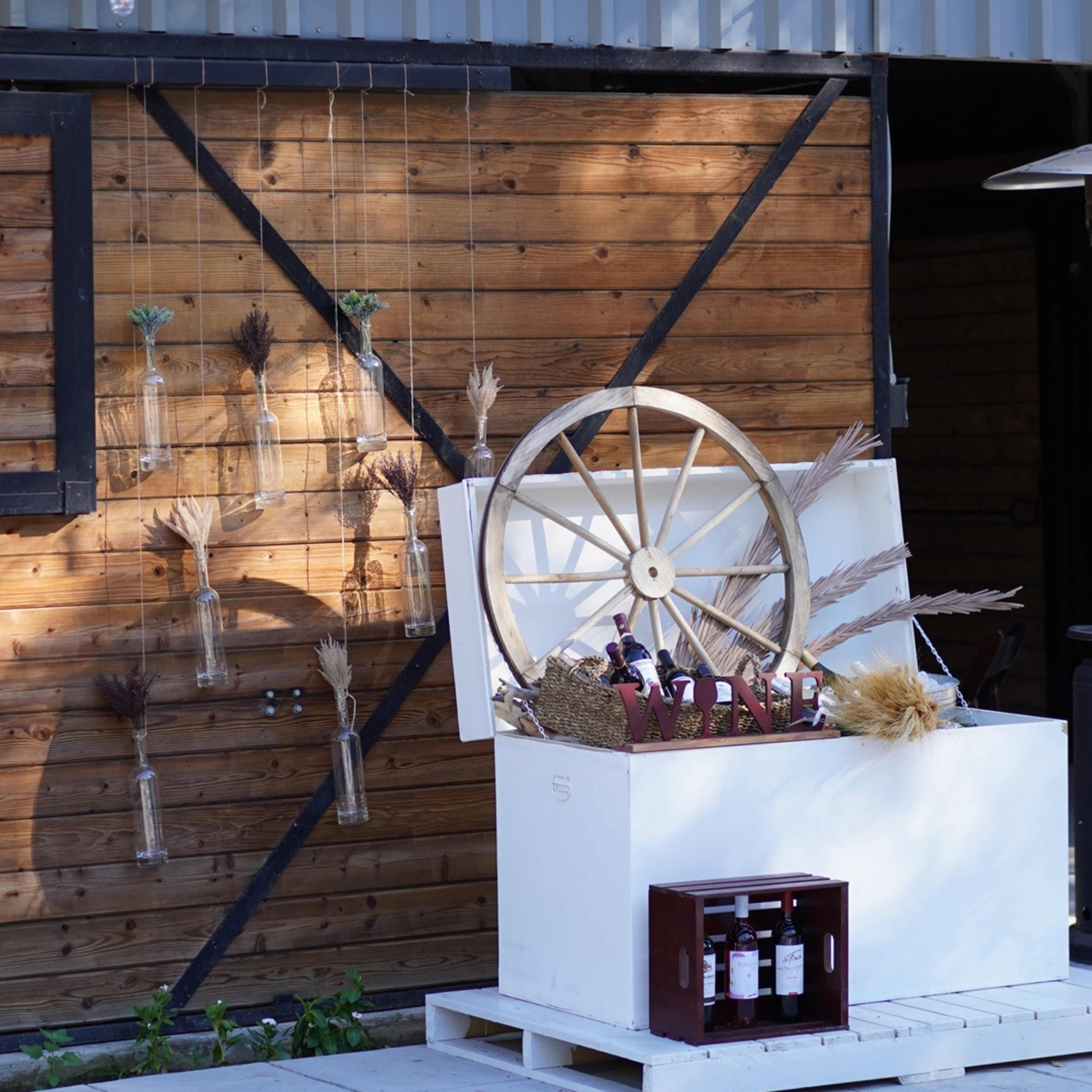 A rustic wooden structure features a decorative display with a large wagon wheel and hanging glass vases, alongside a white crate holding several bottles of wine.