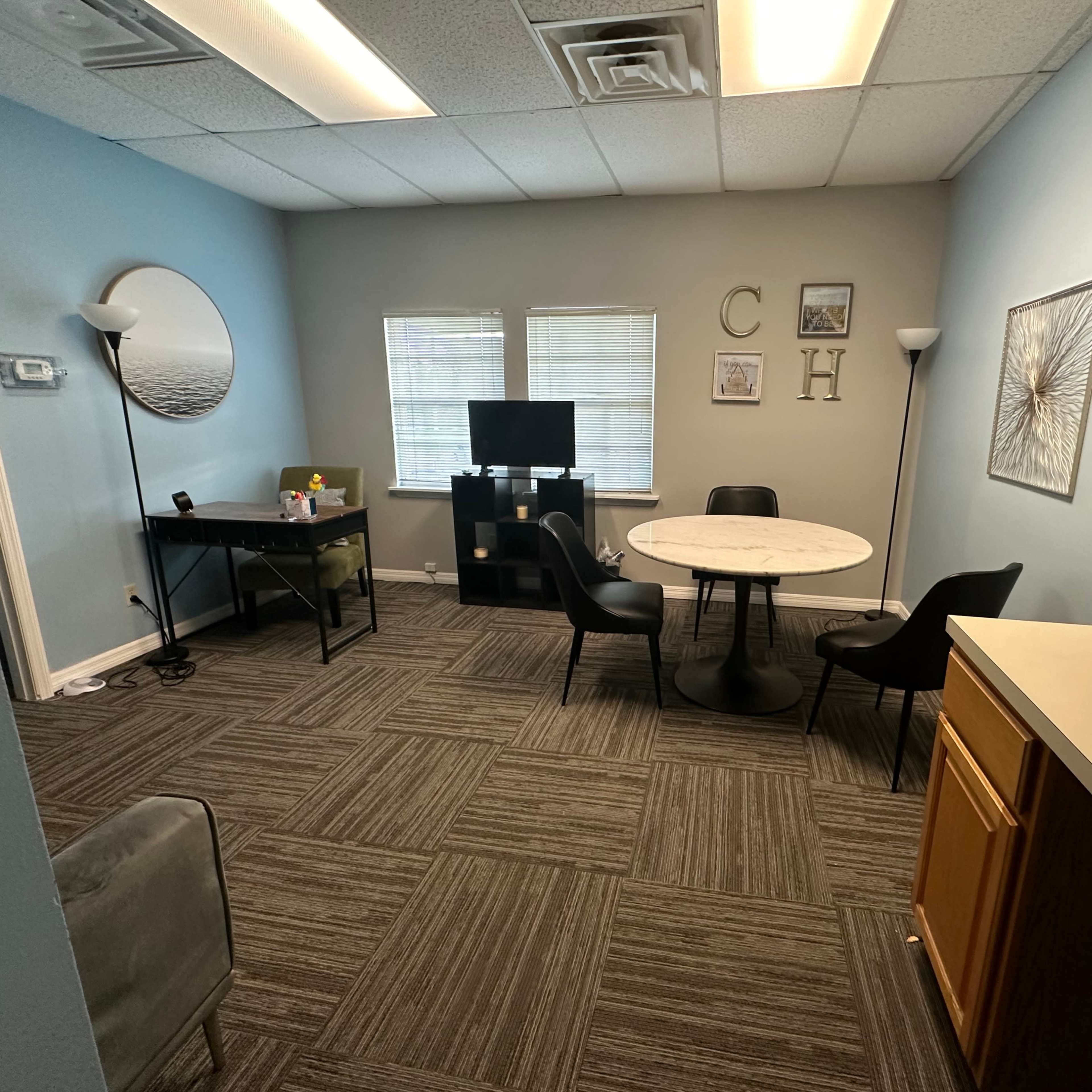 A small office space with a circular table, four chairs, a desk, a TV, and light blue walls.