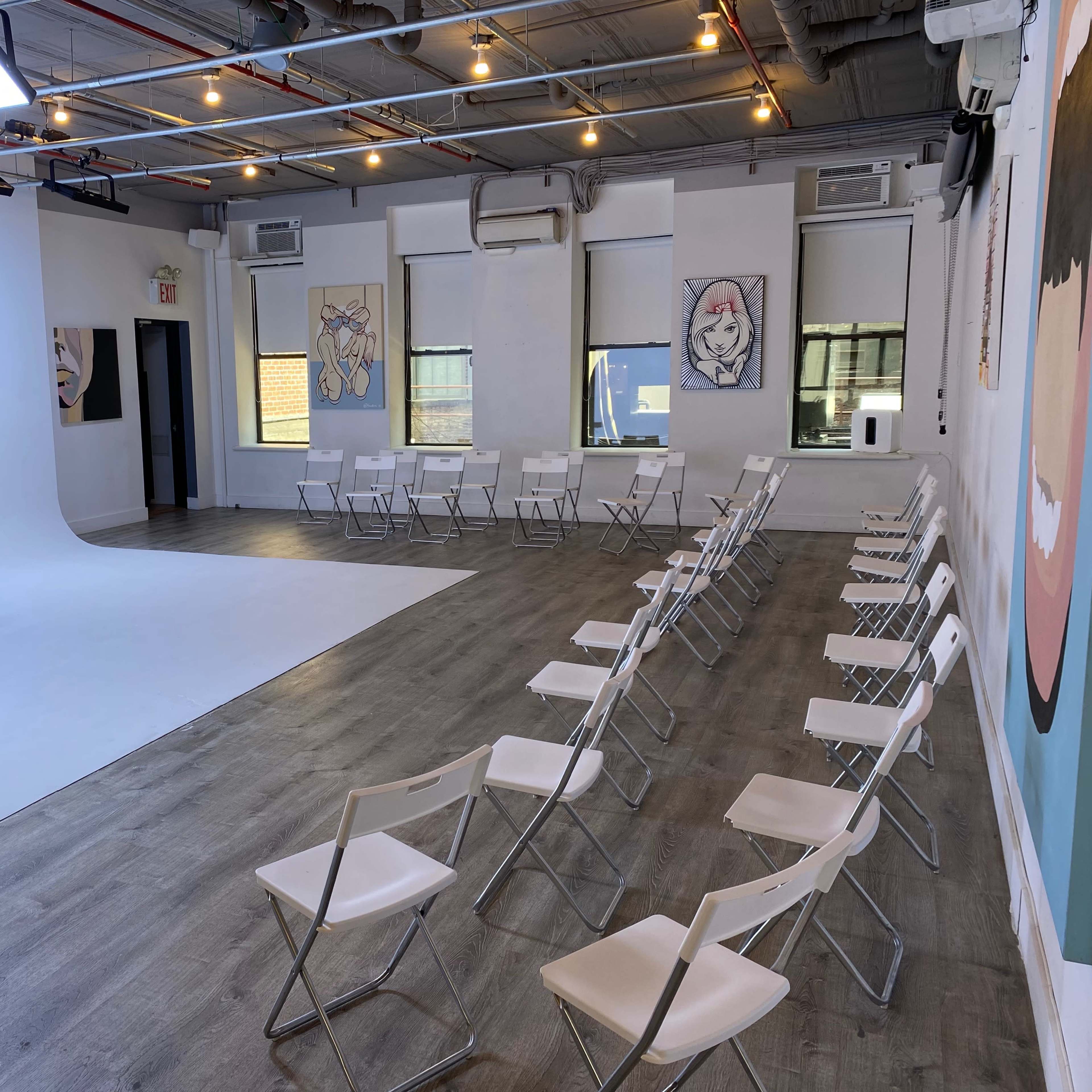 A spacious, well-lit room with white walls, featuring a curved backdrop, two stools on a stage-like area, and rows of white folding chairs arranged for an audience.