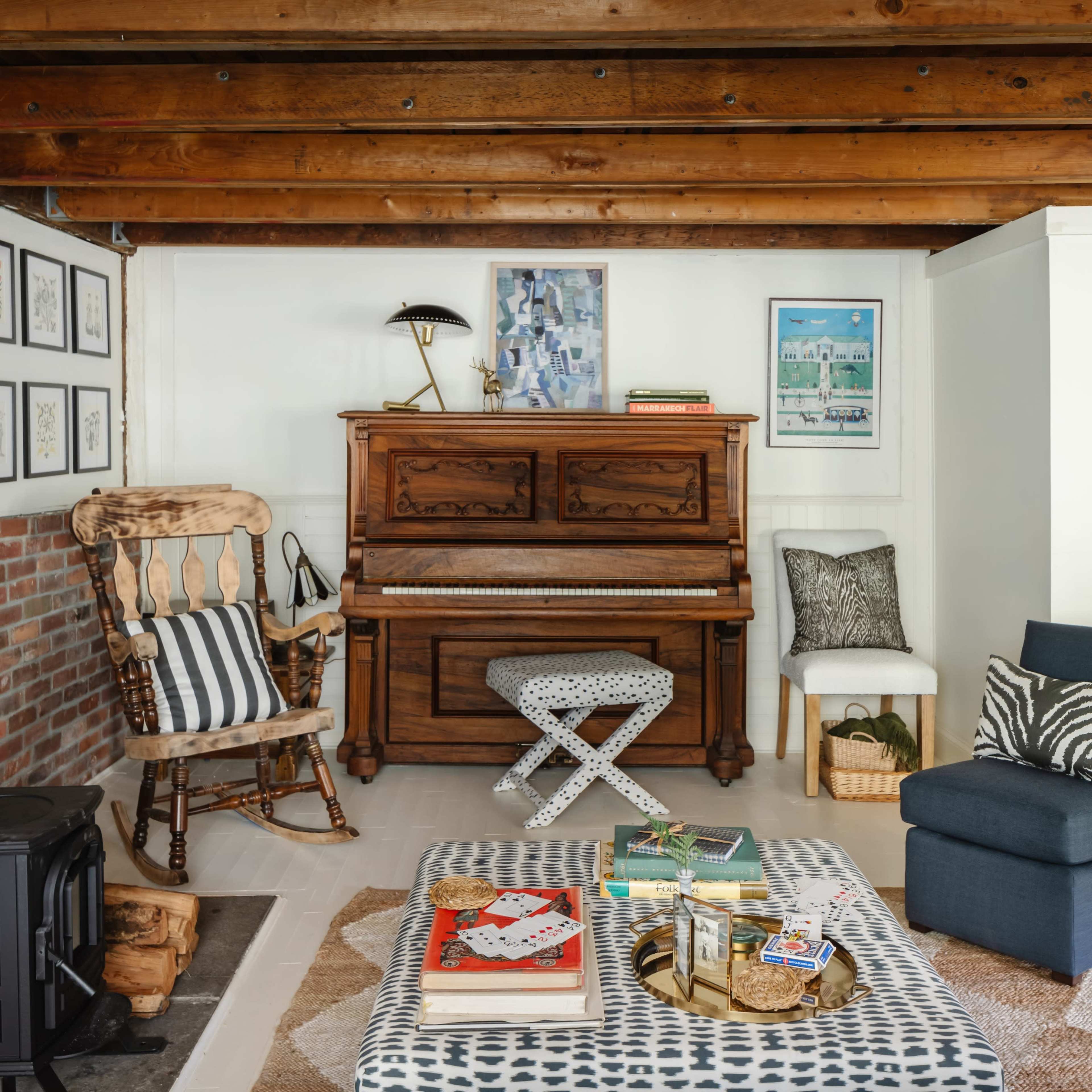 The living room features a wooden piano against a backdrop of exposed beams, with a mix of furniture including a rocking chair, an ottoman, and a coffee table adorned with magazines.