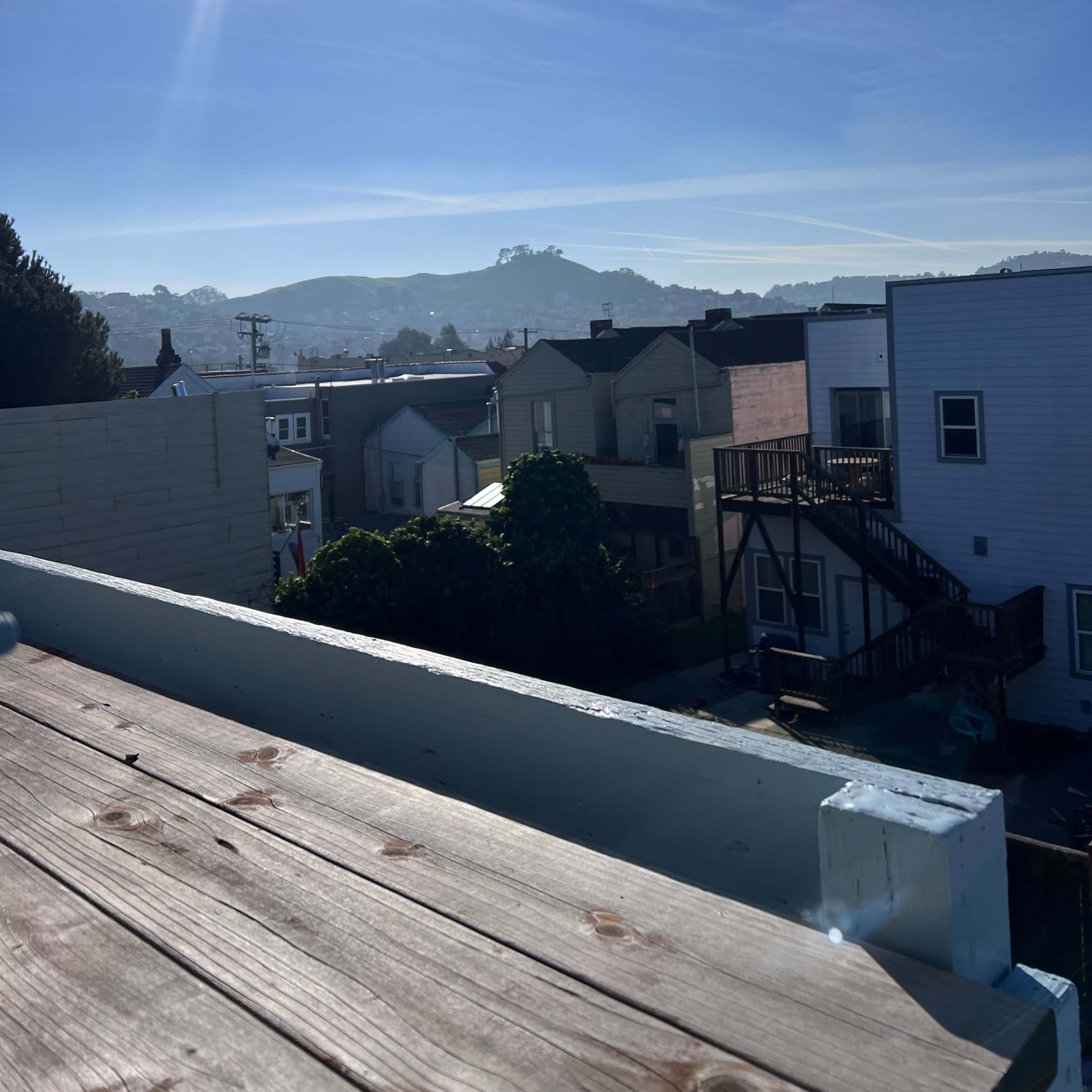 A view from a rooftop, overlooking a neighborhood with several houses and a distant hillside under a clear blue sky.
