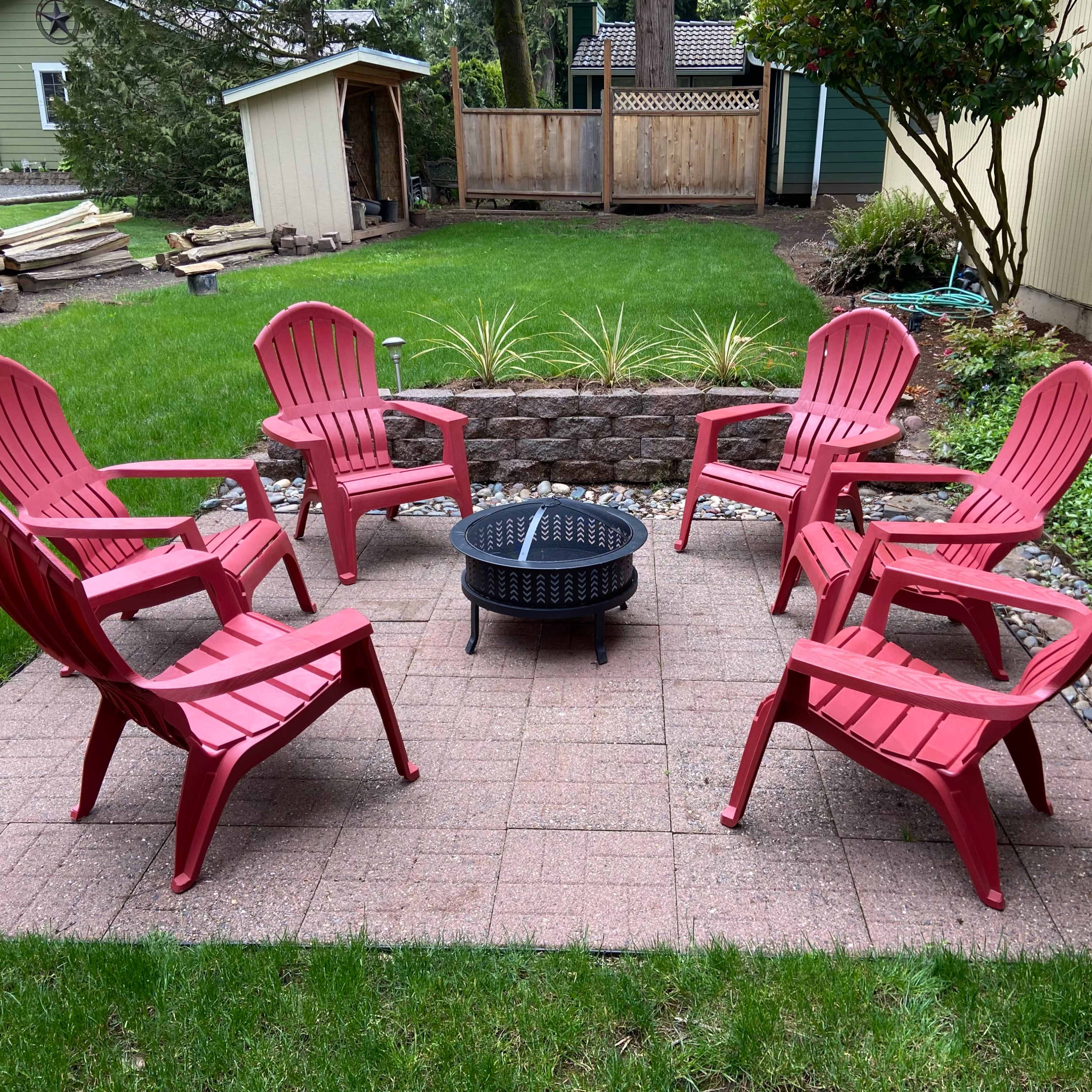 A circular arrangement of six red plastic chairs surrounds a fire pit on a paved patio in a grassy backyard.