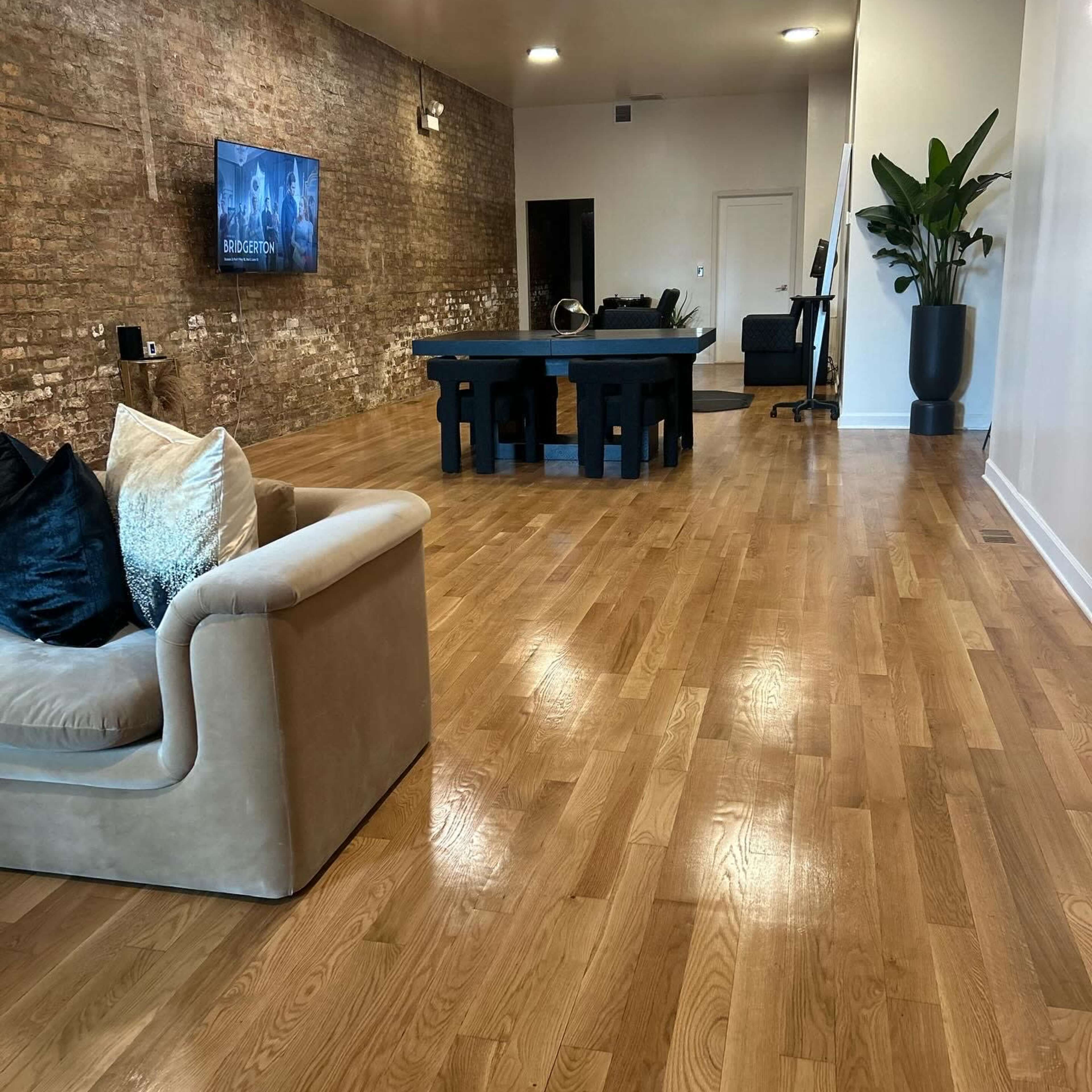 A spacious room with wooden flooring, a sectional sofa, a large dining table, and a potted plant, along with a wall-mounted TV and an office chair in the background.