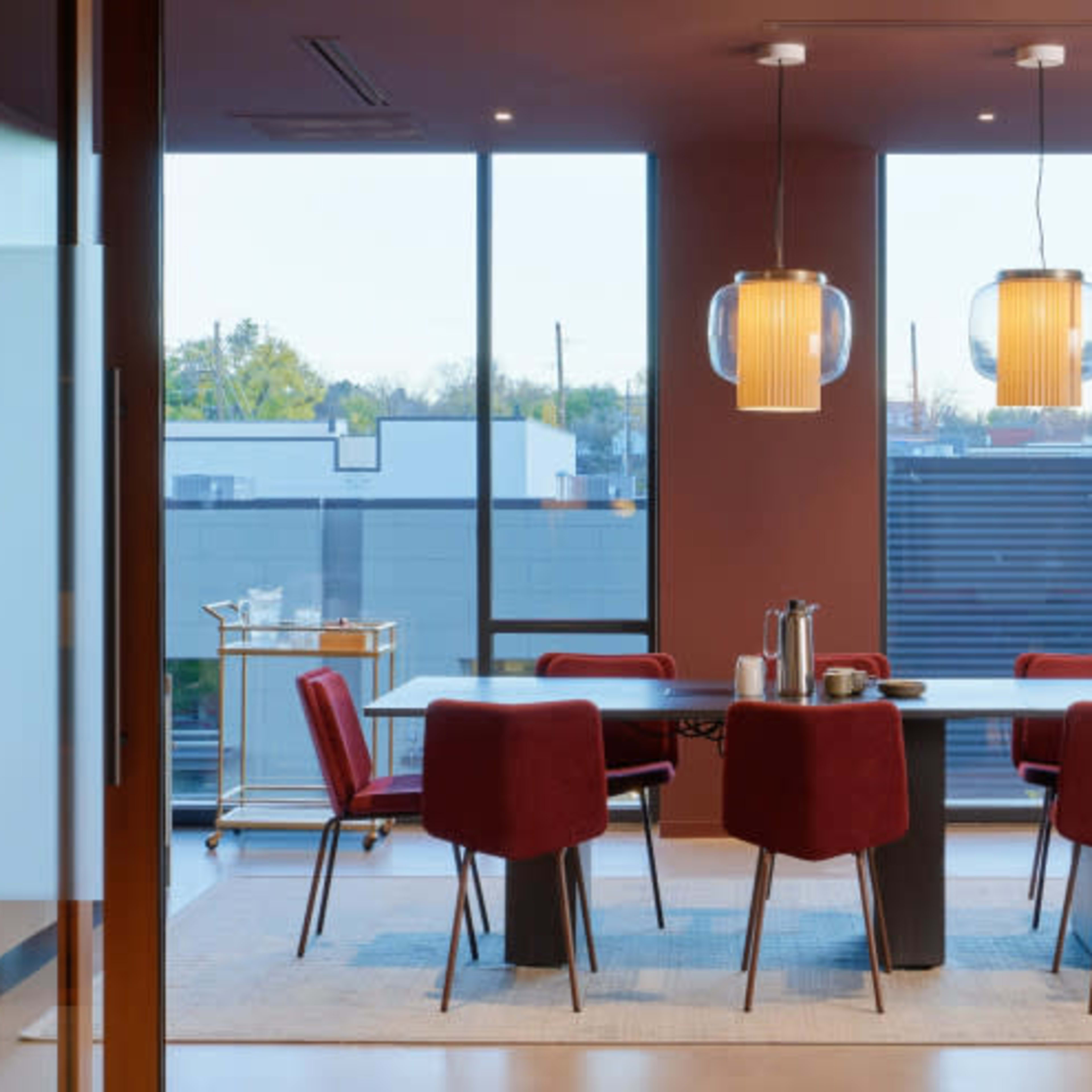 A modern dining area features a large table surrounded by red chairs, with pendant lighting and large windows providing natural light.