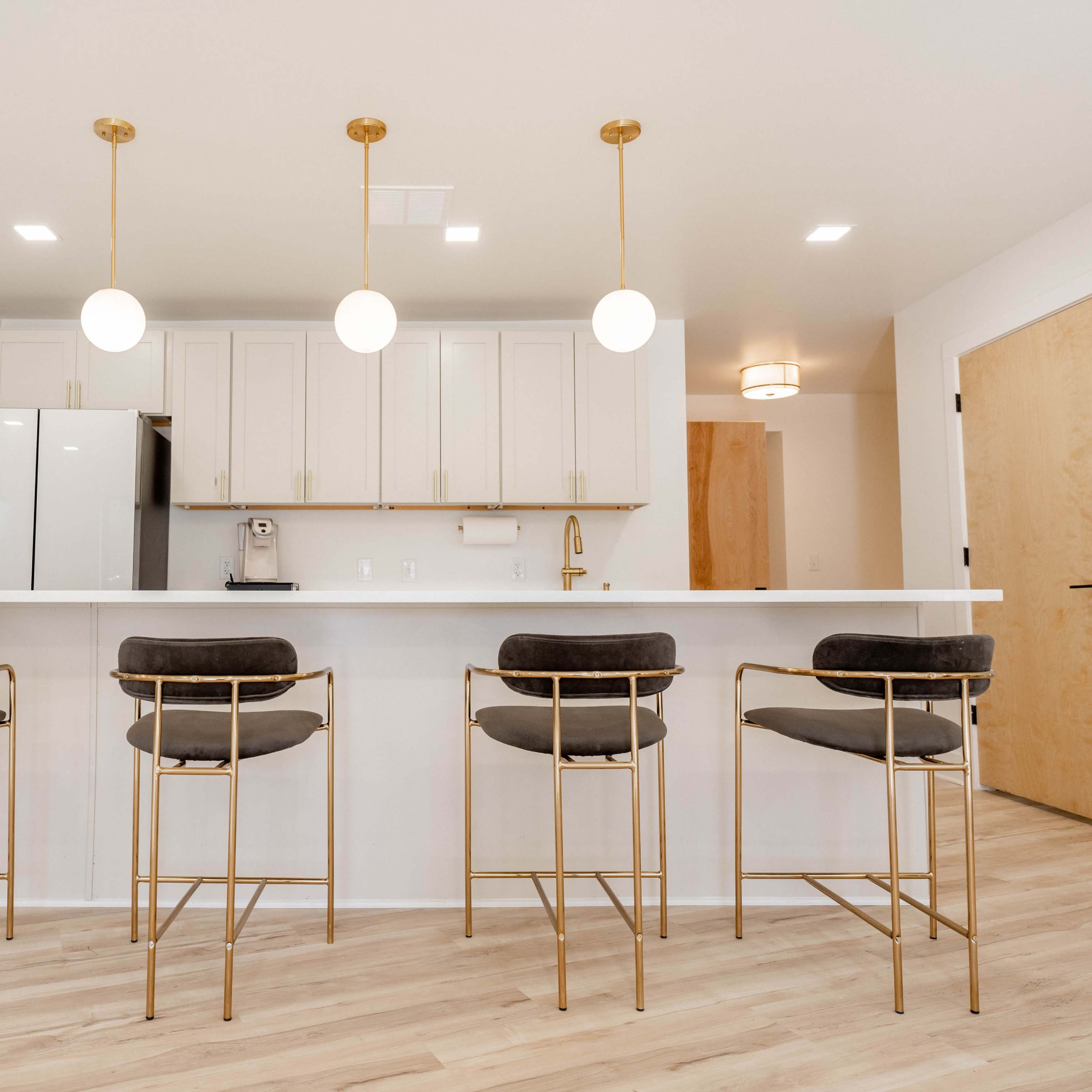 A modern kitchen with a white countertop, four gold-framed bar stools, and pendant lights hanging above.