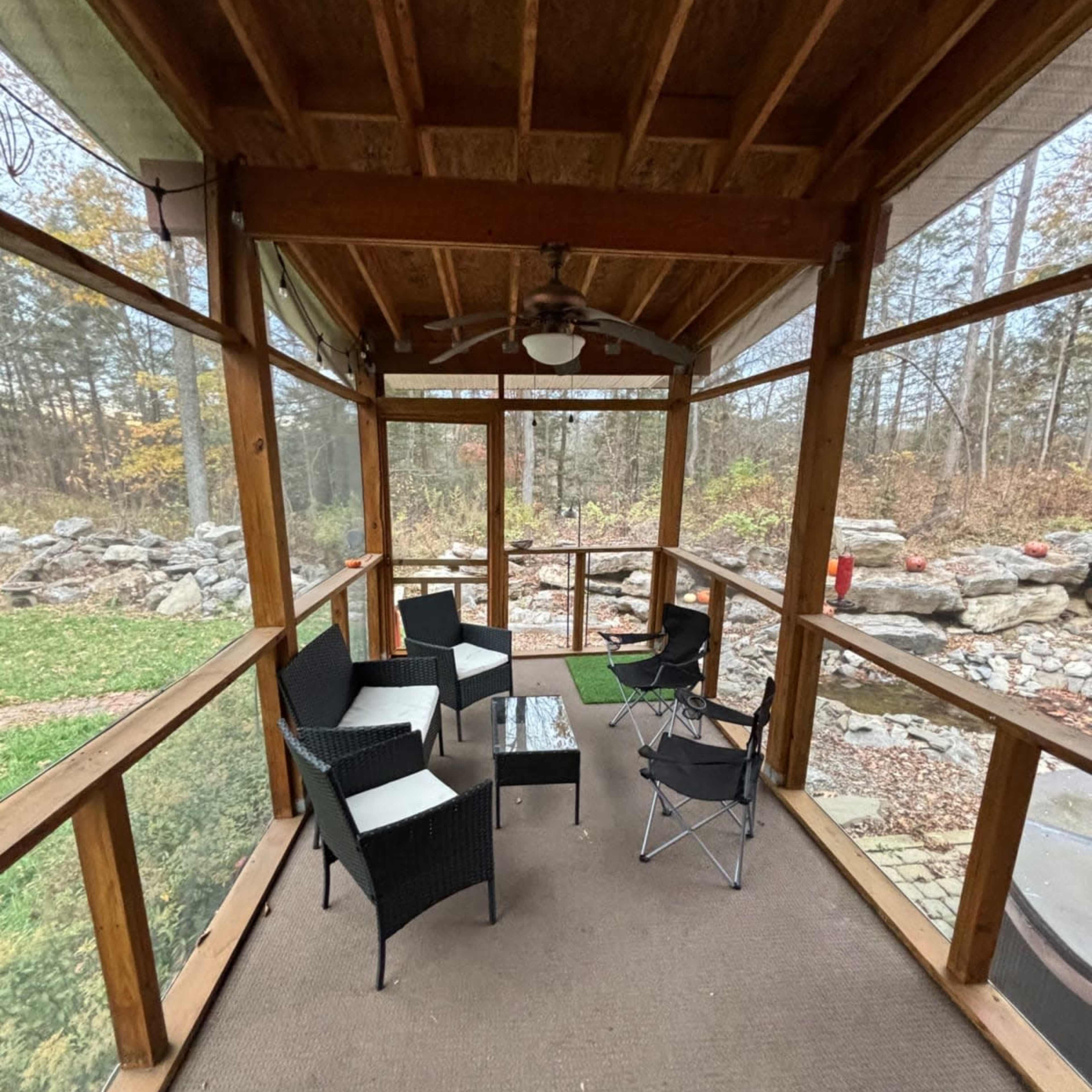 A screened porch with several chairs and a small table, surrounded by trees and a stone wall in the background.