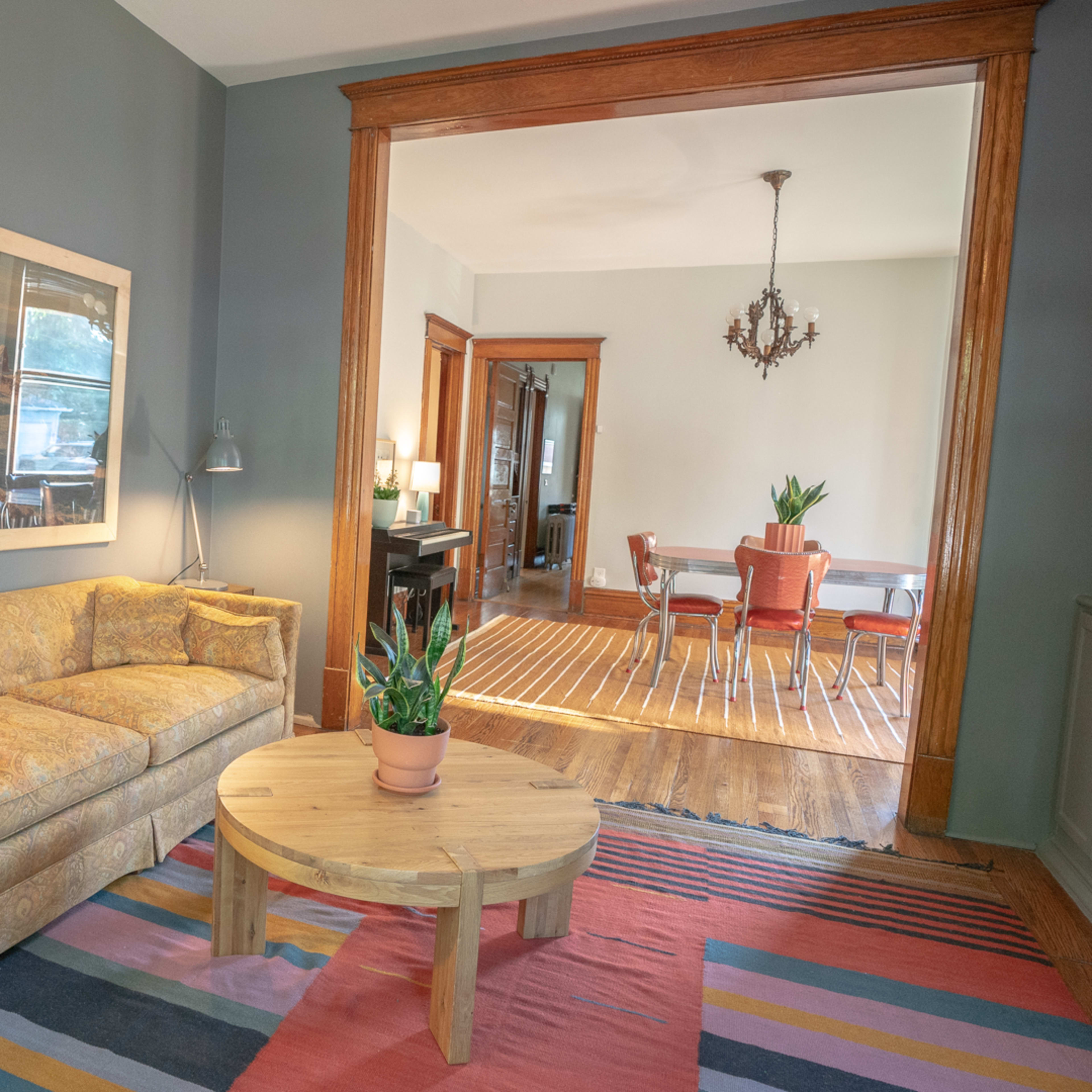 A living room with a patterned sofa, a round wooden coffee table, and a view into a dining area featuring a chandelier and additional seating.