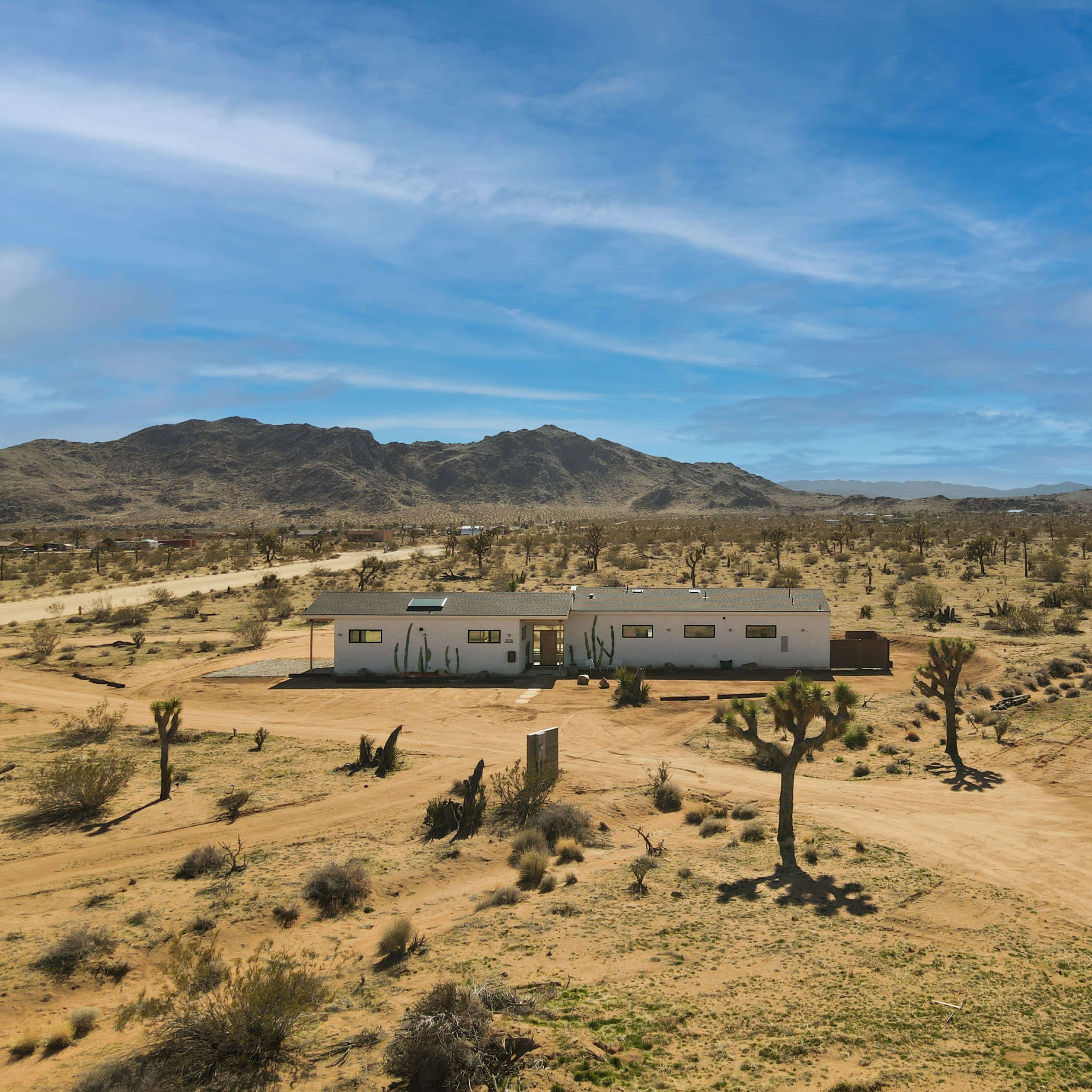 A white house stands in a desert landscape surrounded by sparse vegetation and mountains in the background.