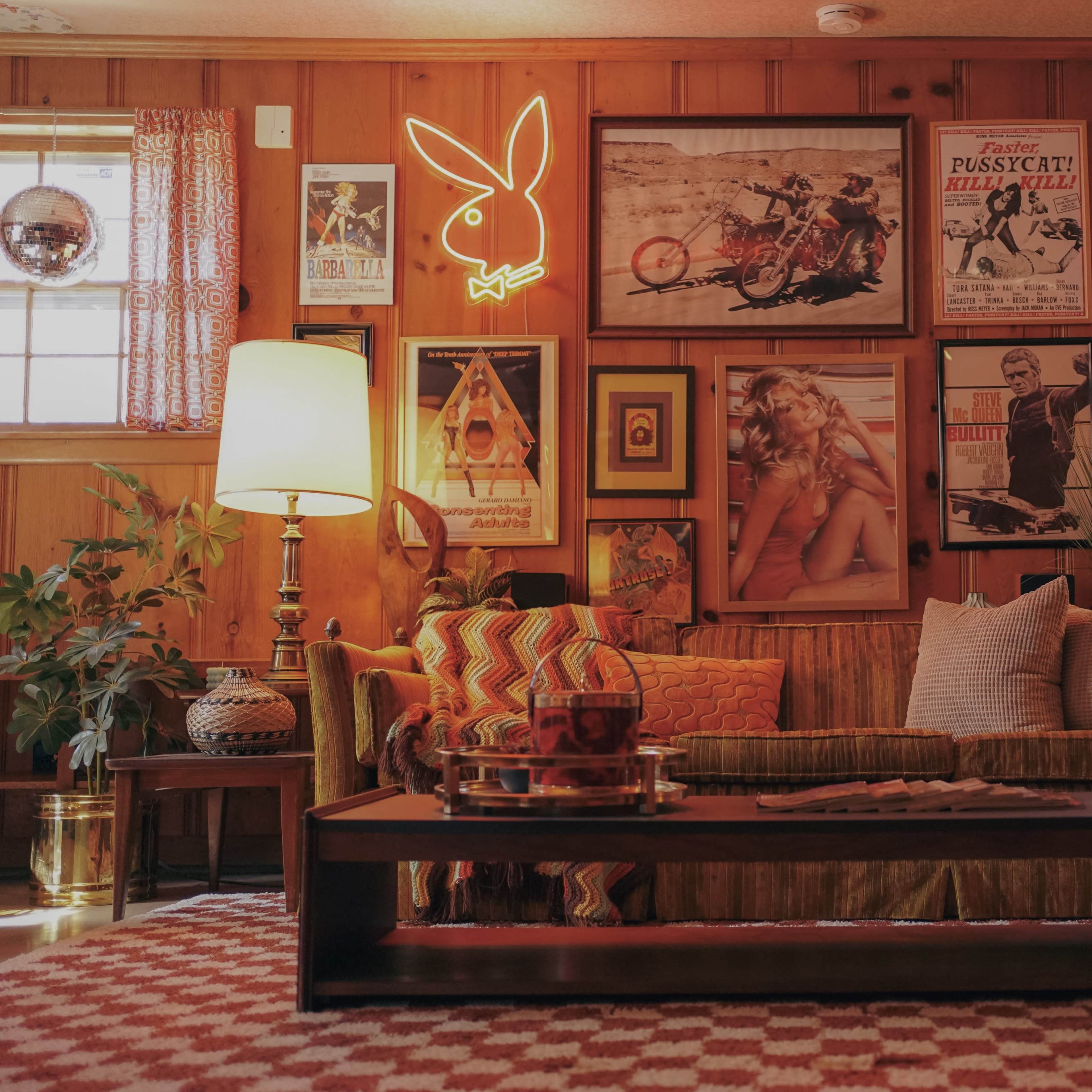 A cozy living room with wooden paneling, vintage posters on the walls, a neon Playboy sign, and a sofa with decorative cushions in front of a coffee table.