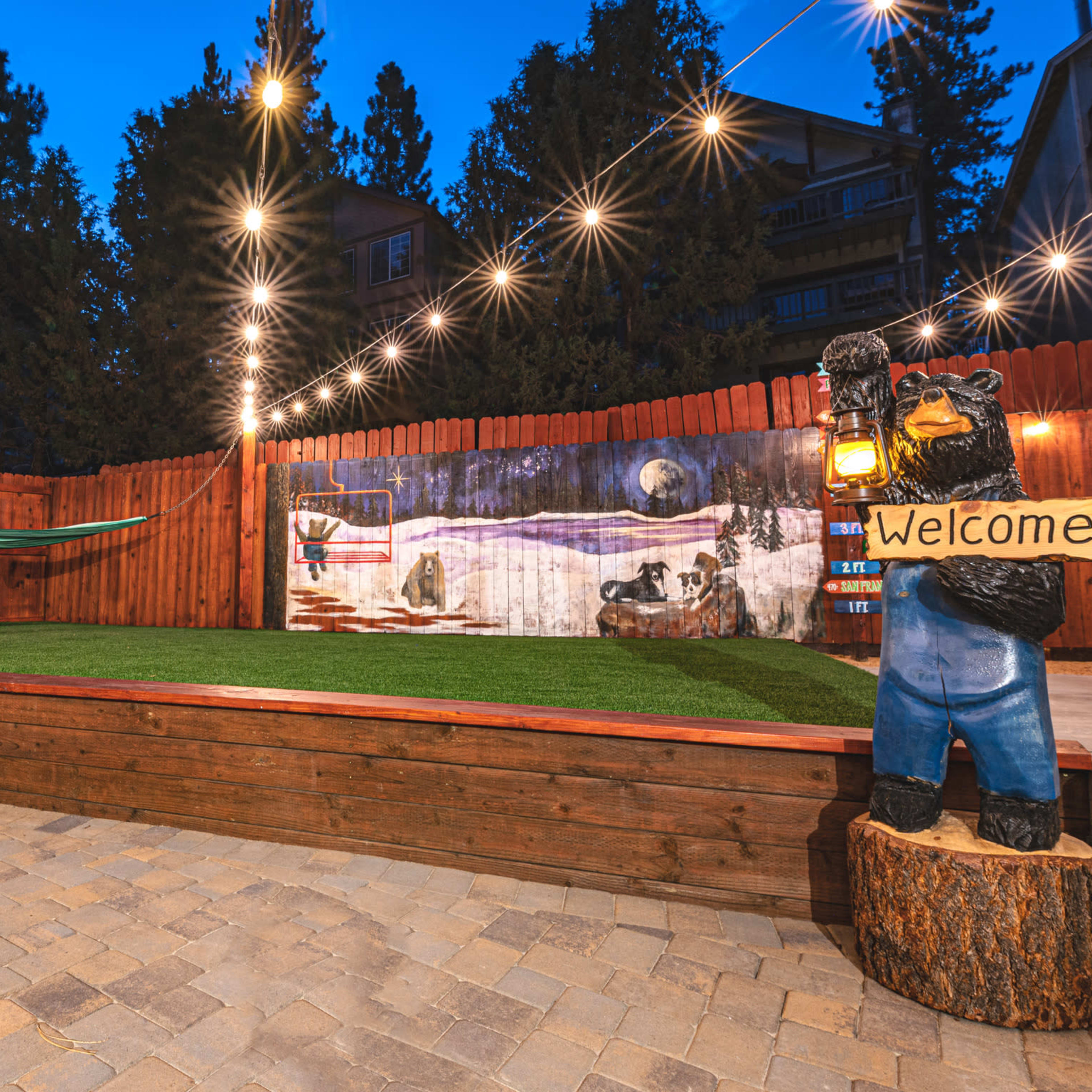 A landscaped backyard with a wooden deck, a hammock, a mural of a winter scene, string lights, and a bear statue holding a "Welcome" sign.