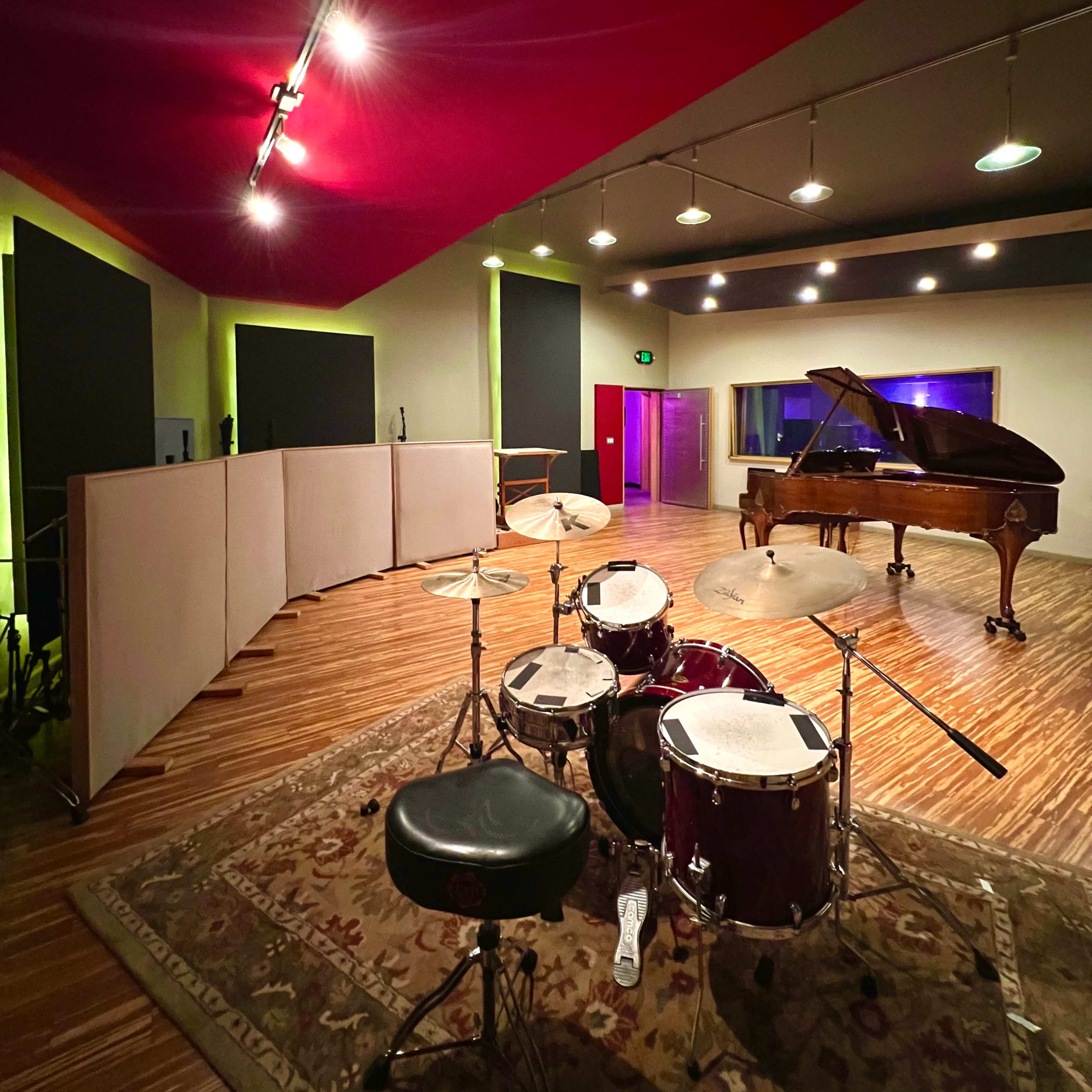 A music studio featuring a drum set in the foreground and a grand piano positioned against a wall, with sound panels installed around the room.