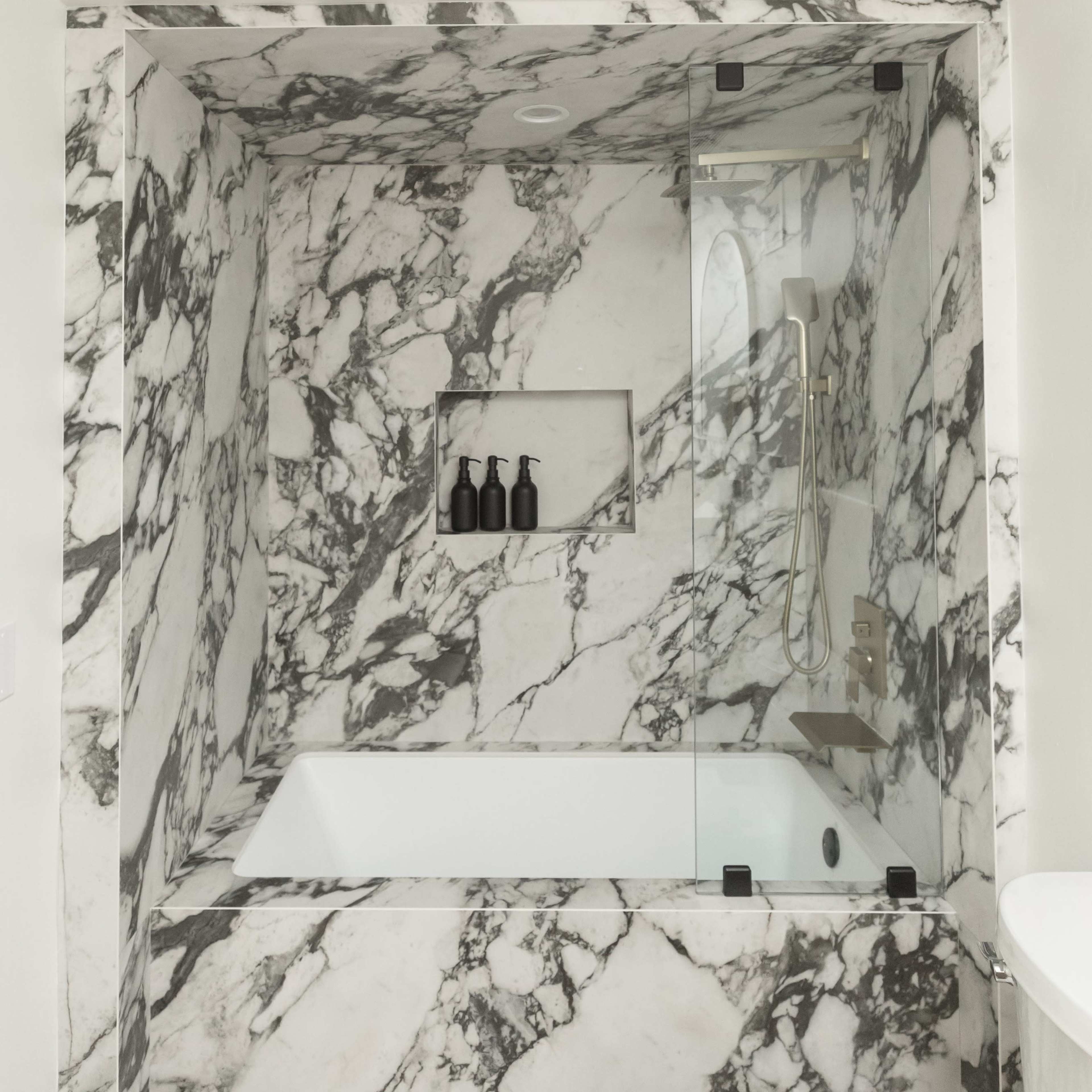 A modern bathroom featuring a marble-clad bathtub enclosure with a glass shower partition and a built-in shelf for toiletries.