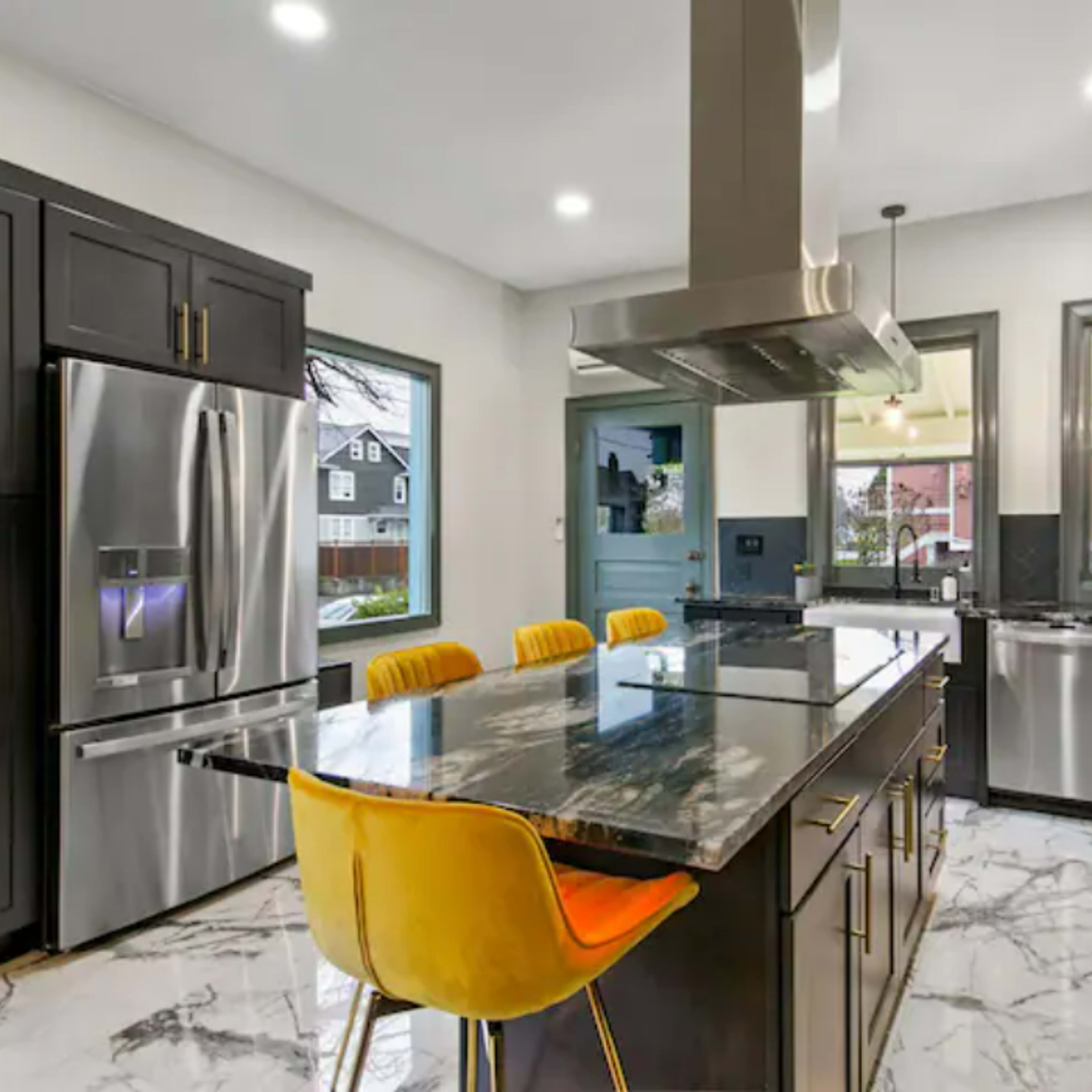 A modern kitchen with dark cabinetry, stainless steel appliances, and a large island featuring yellow stools on a marble floor.