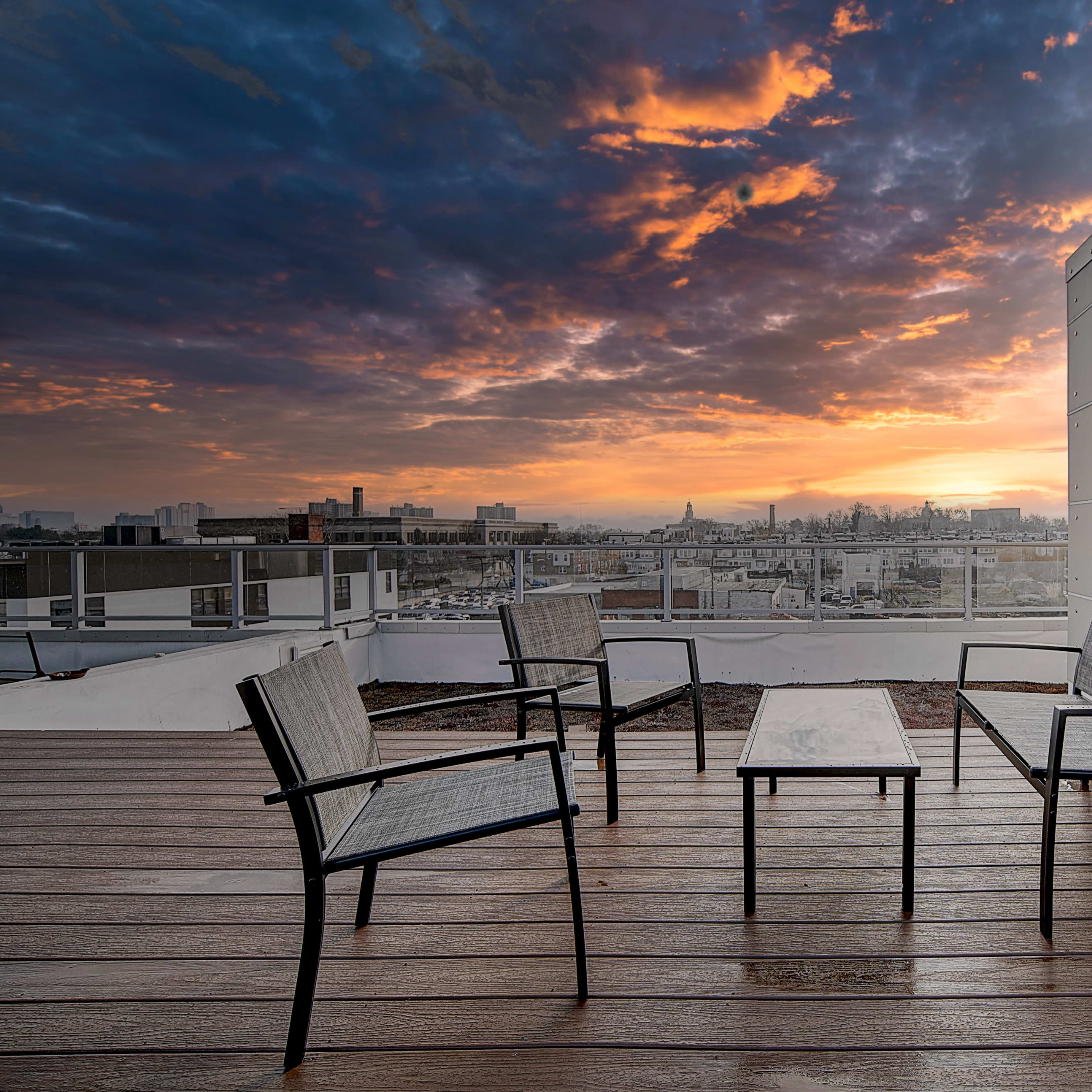 A rooftop patio with four chairs and a table, overlooking a city skyline at sunset.