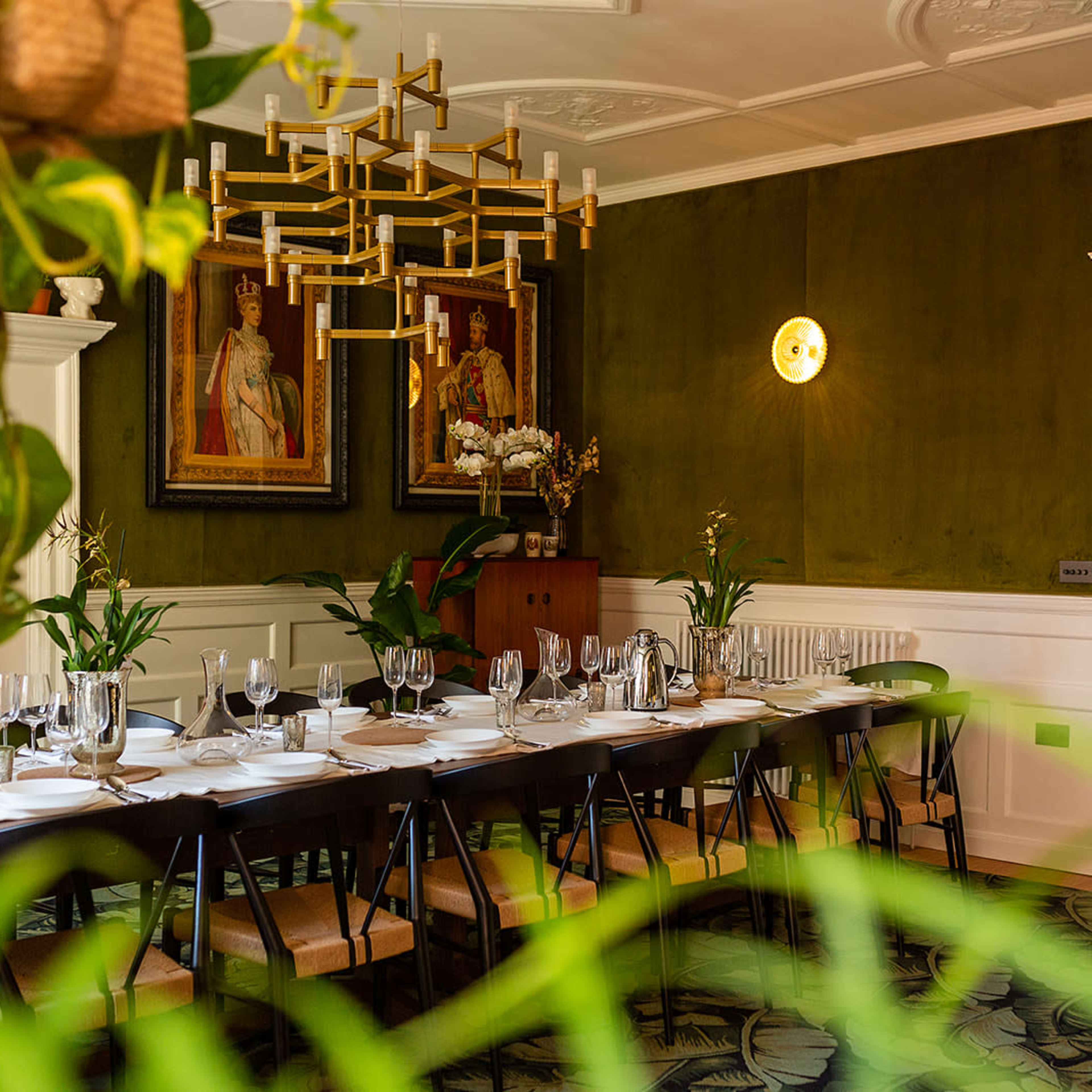 A long dining table is set with tableware in a room featuring green walls, chandeliers, and framed portraits.