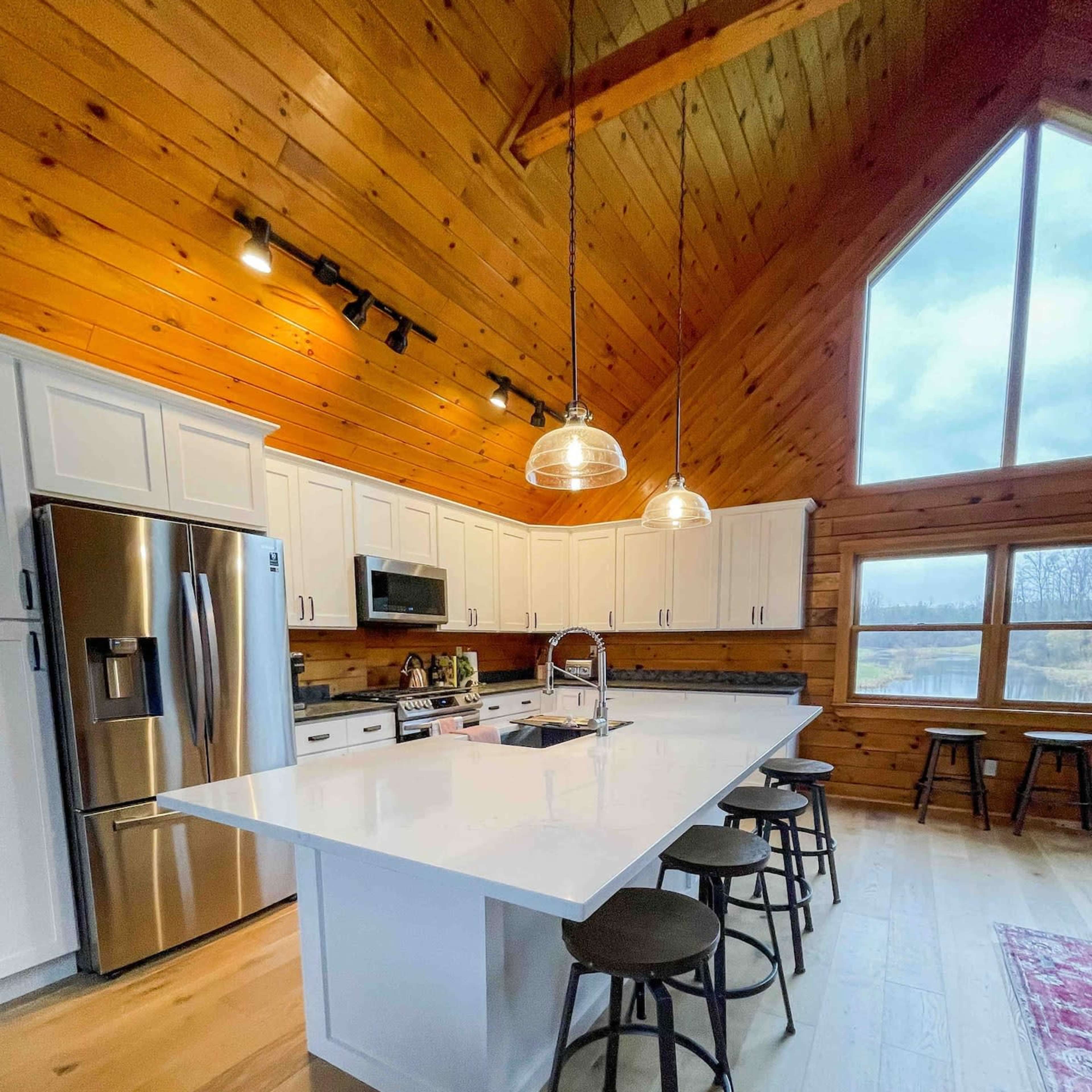 A modern kitchen with a large white island, stainless steel appliances, and wooden walls under a vaulted ceiling.