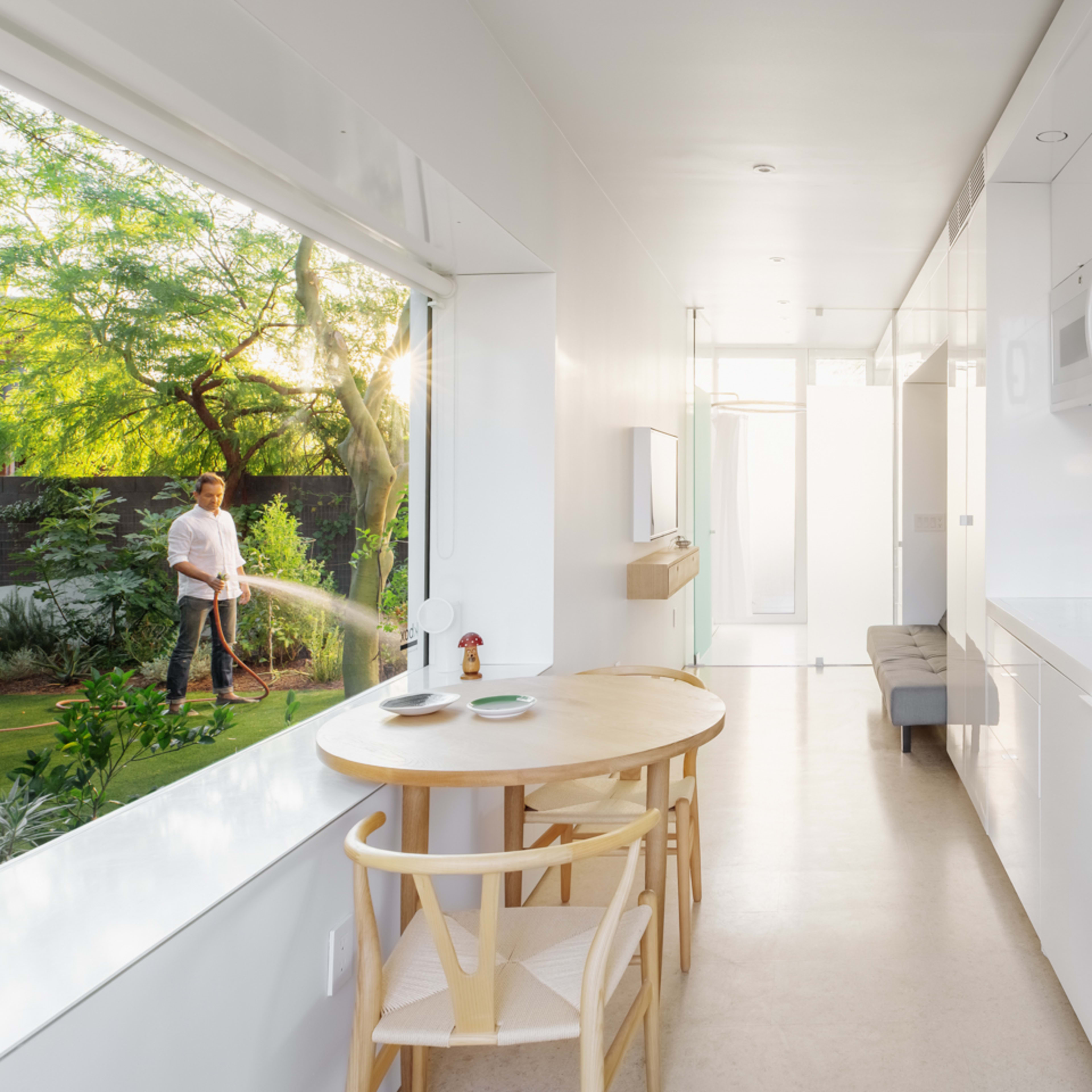 A bright, modern kitchen features a dining table and a view of a person watering plants in a lush garden outside.