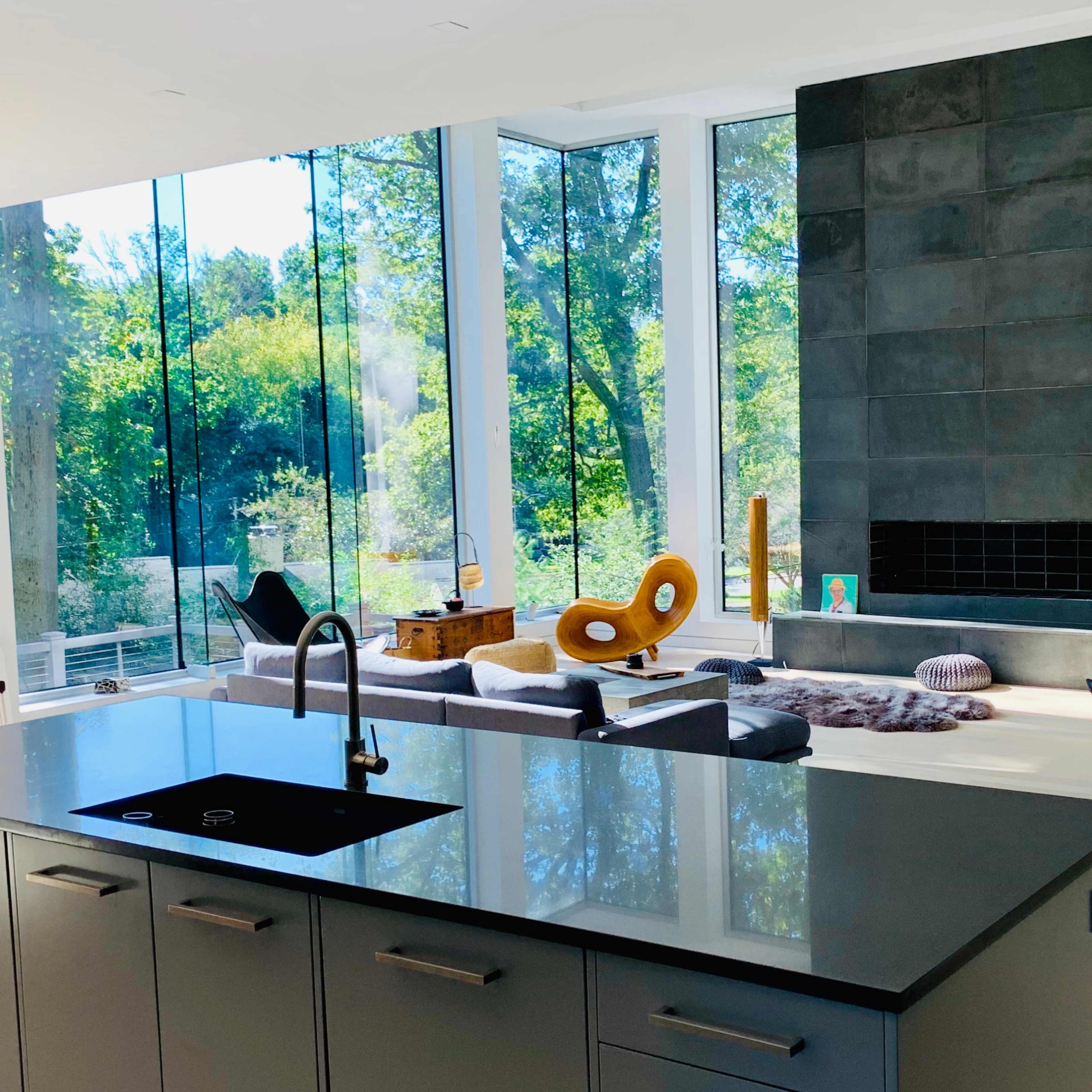 A modern living space features a kitchen with a black countertop, large windows overlooking a wooded area, and a cozy seating arrangement near a stone fireplace.