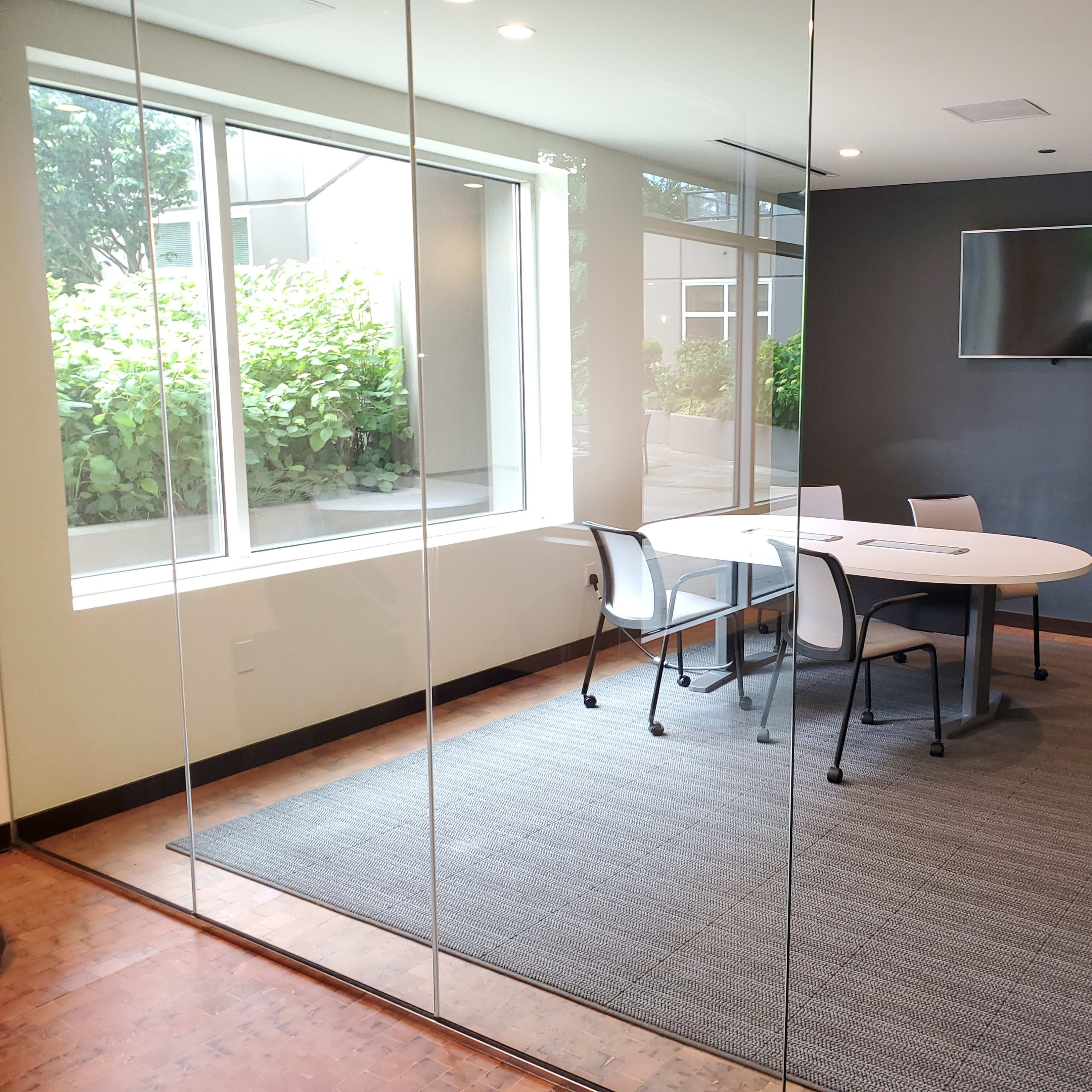 A modern conference room with a round table, four chairs, a large television on the wall, and a green view outside through a glass wall.