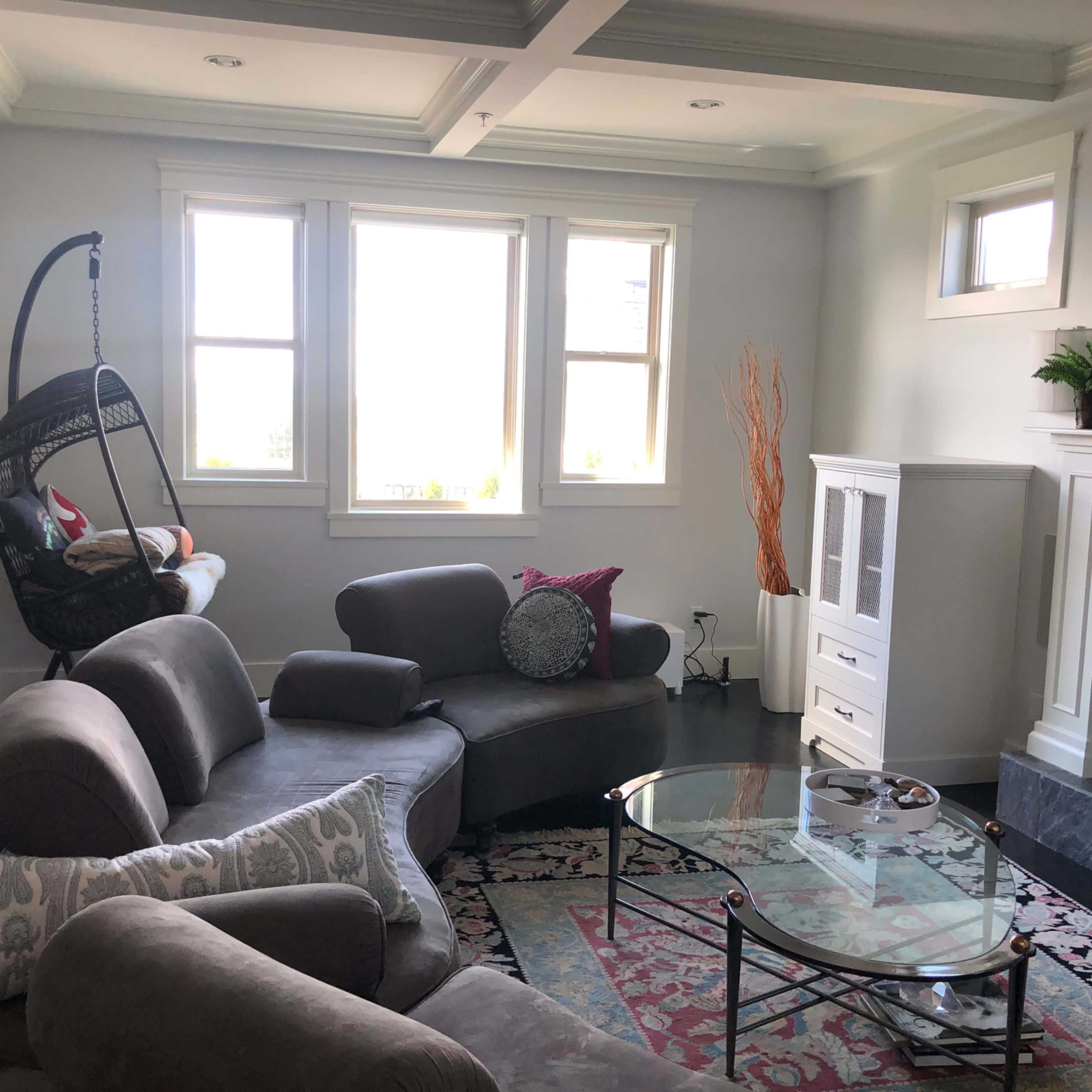 The living room features a curved gray sectional sofa, a glass coffee table, and large windows that allow natural light to enter the space.
