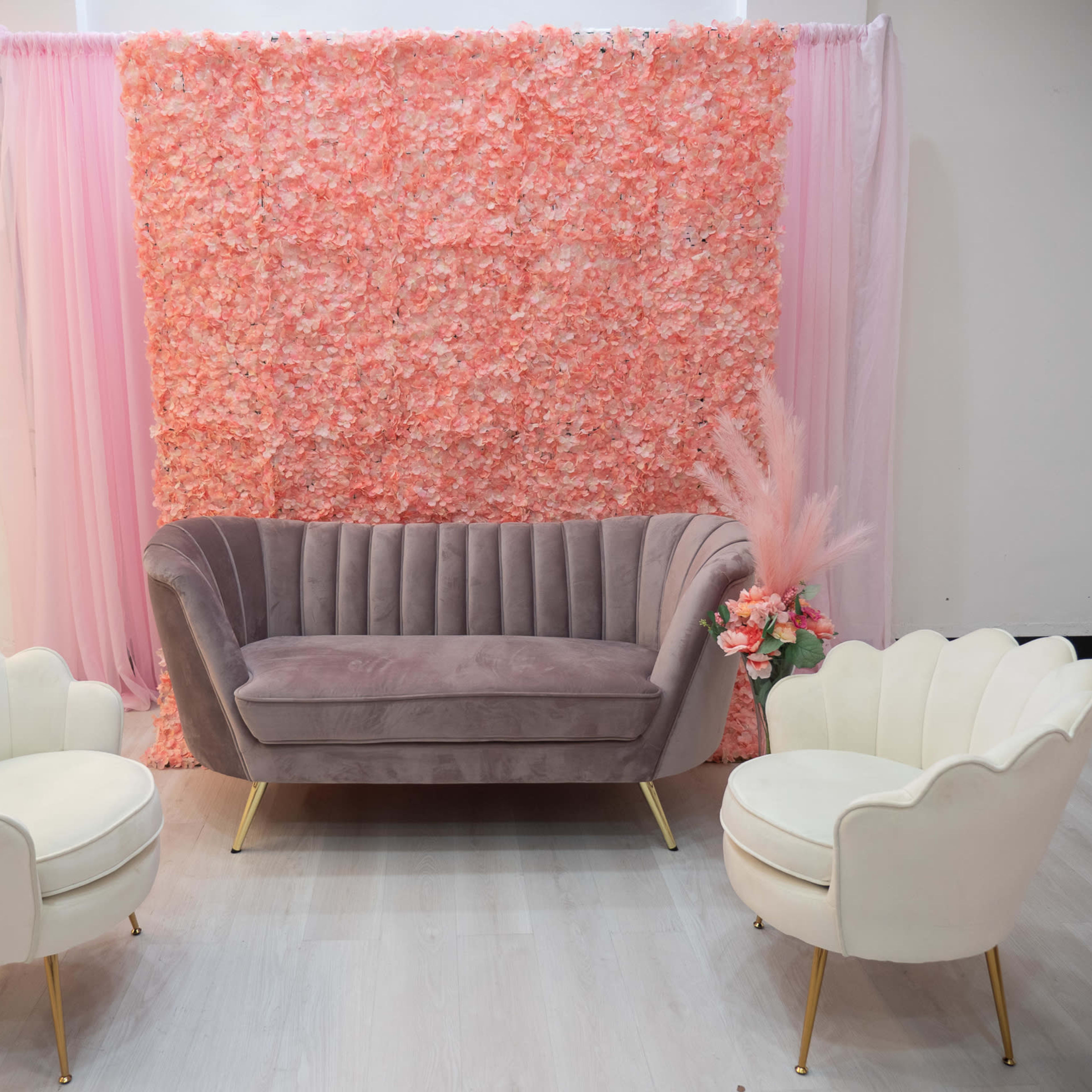 A modern seating arrangement featuring a grey velvet sofa and two white armchairs, set against a floral pink backdrop.