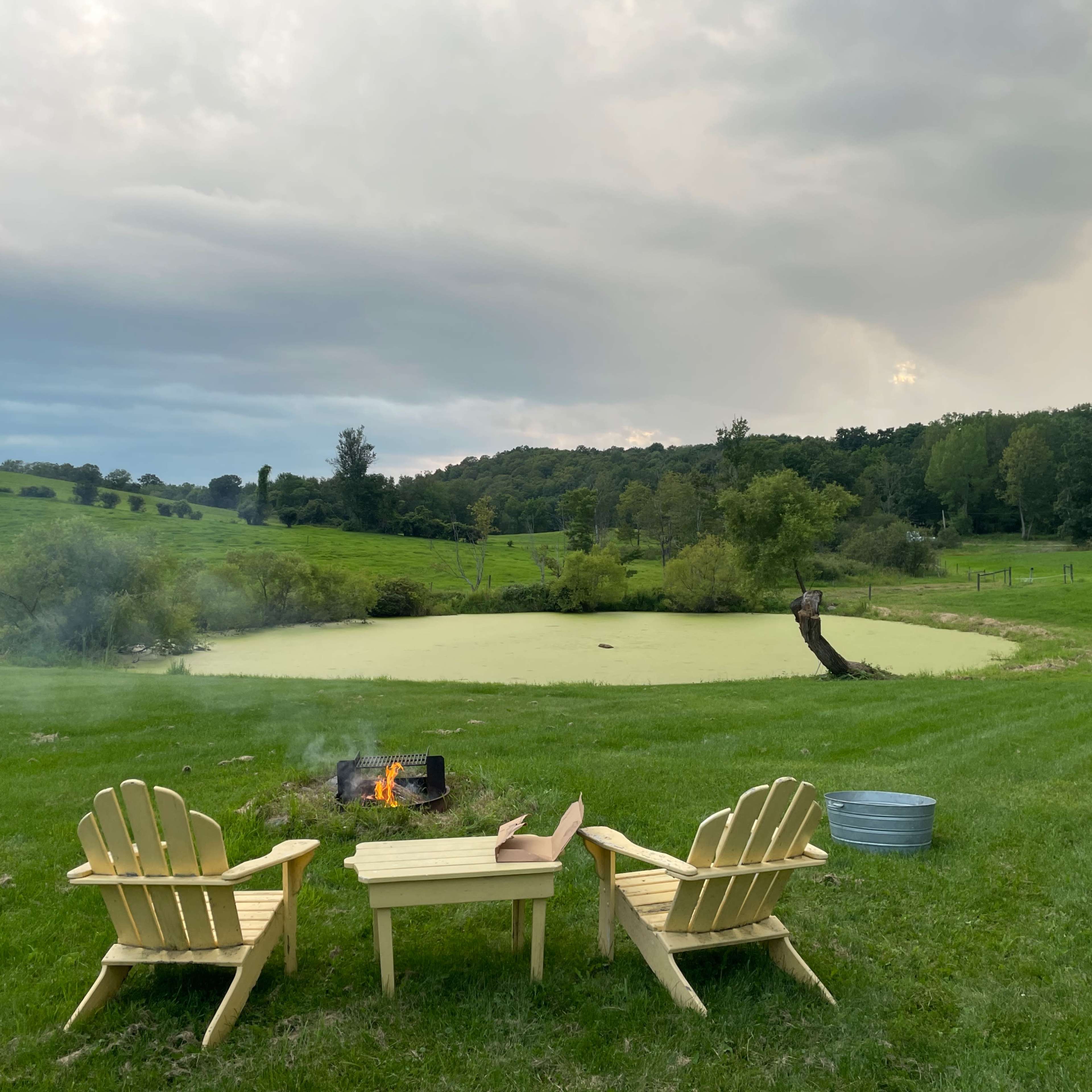 Two wooden chairs and a small table sit beside a pond covered in green algae, with a fire pit burning in front of them on a grassy field.