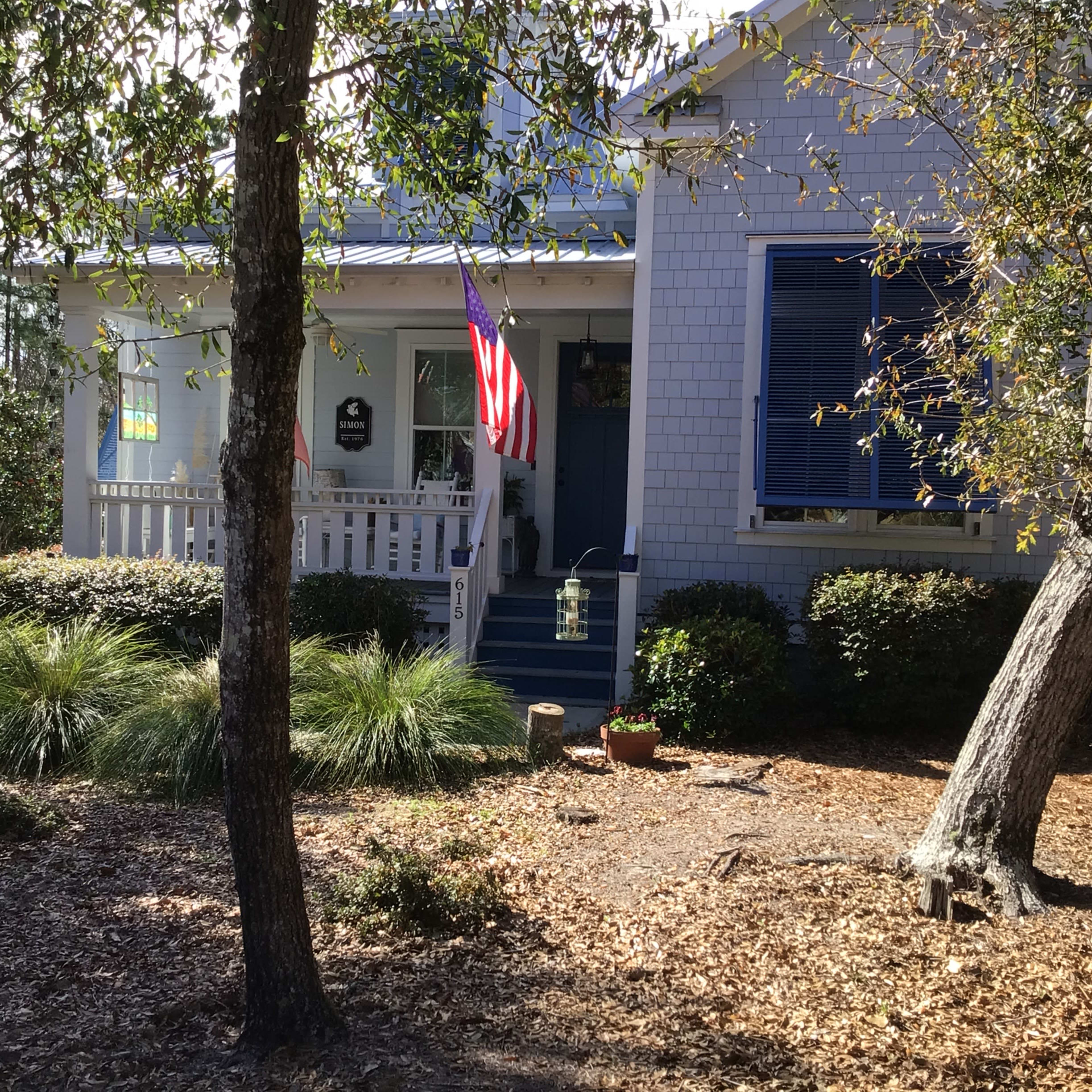 A white house with blue shutters and an American flag displayed on the front porch is surrounded by greenery and mulch.