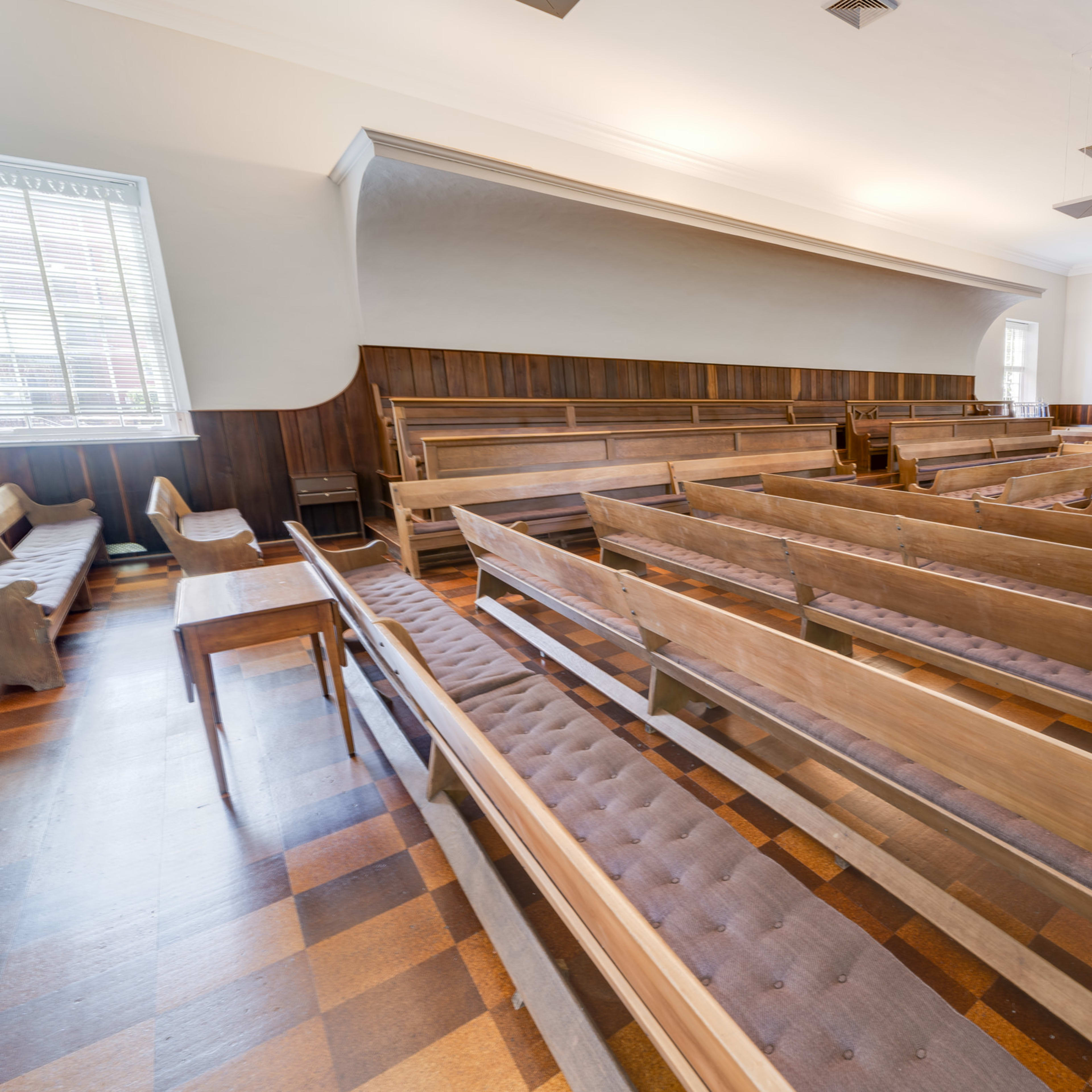 A spacious interior of a room with wooden benches arranged in rows on a checkered floor.