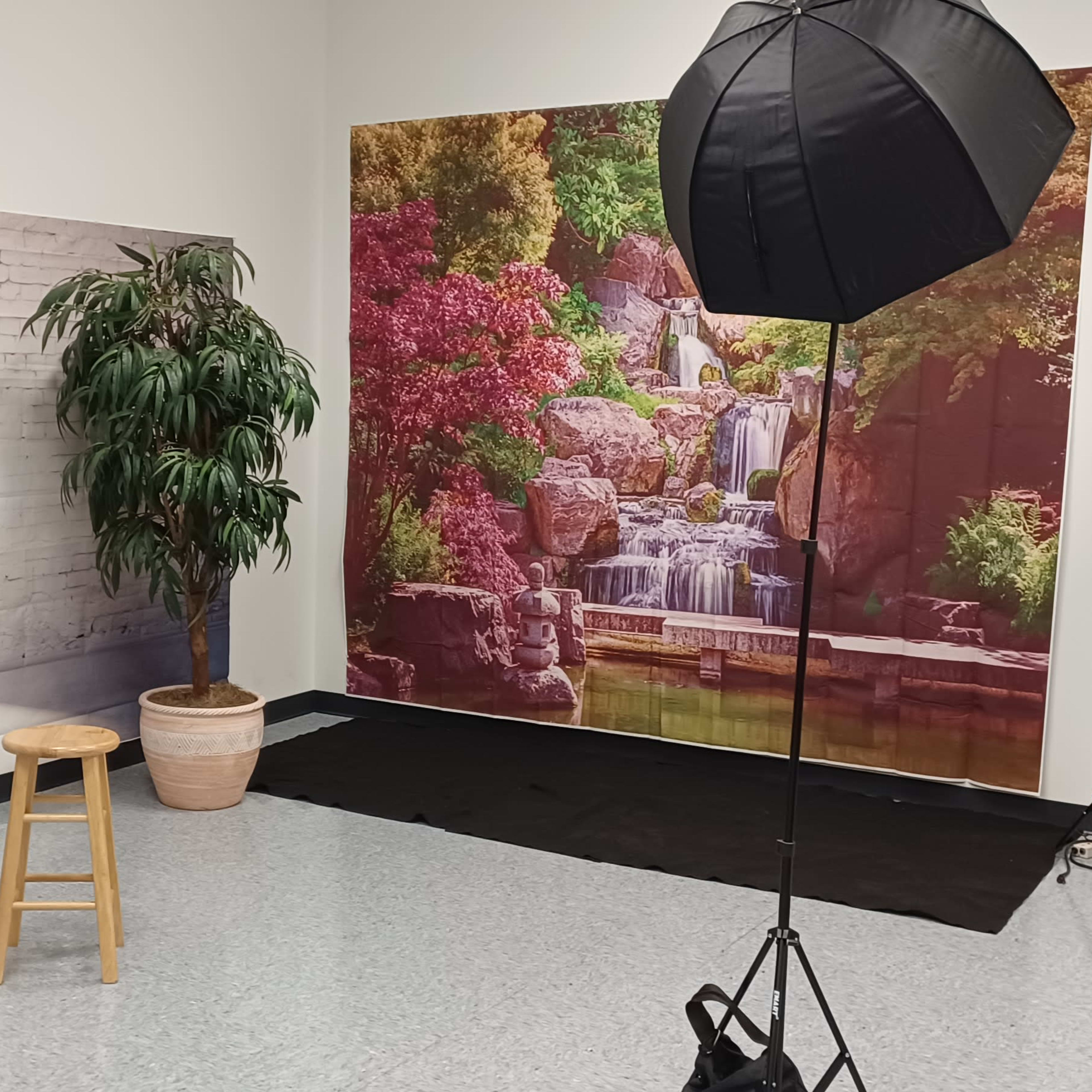 A photography studio setup with two softbox lights, a wooden stool, and colorful backdrop images depicting a waterfall scene and an interior view.
