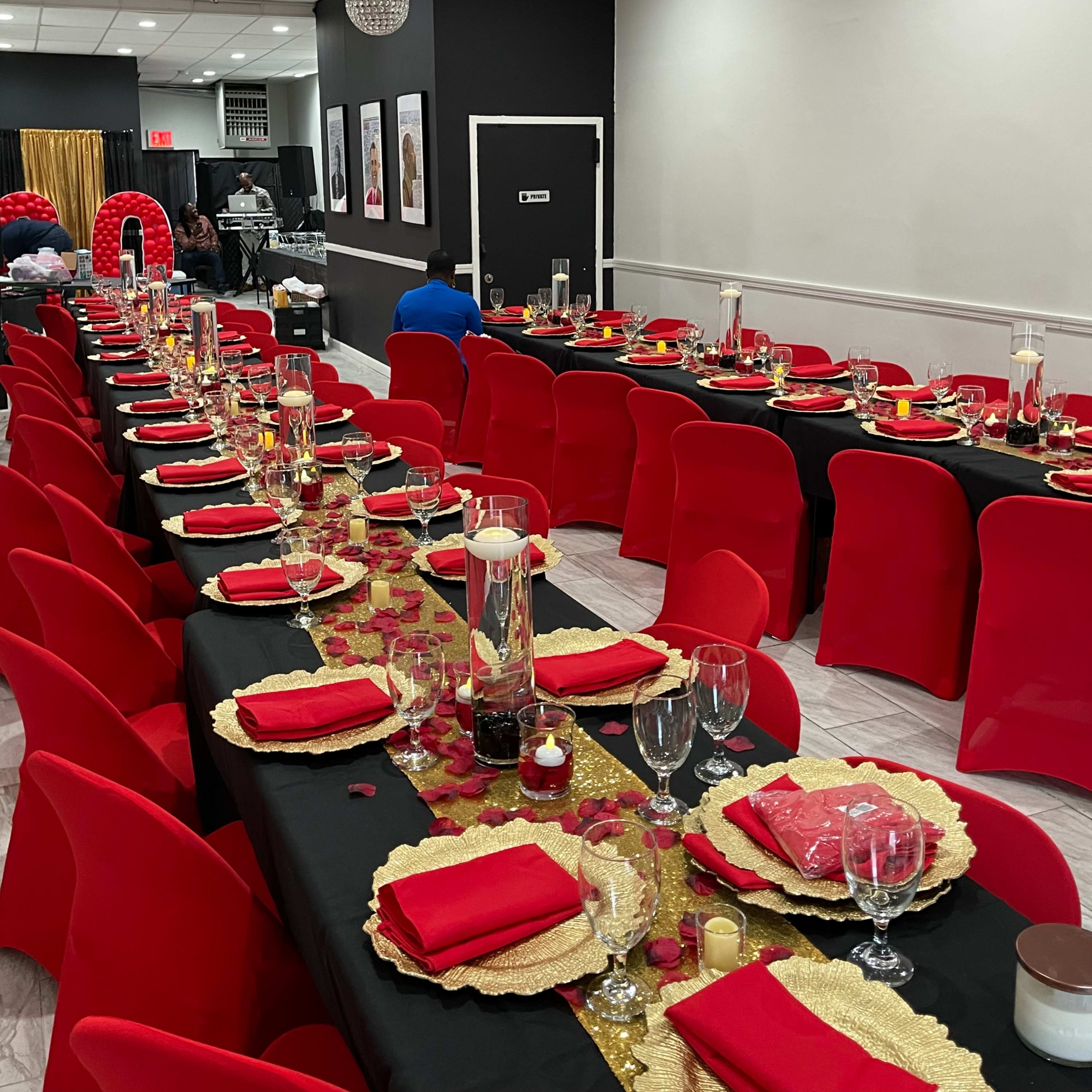 A festive dining setup with long tables covered in black cloth, red chairs, and decorative centerpieces featuring candles and gold accents.