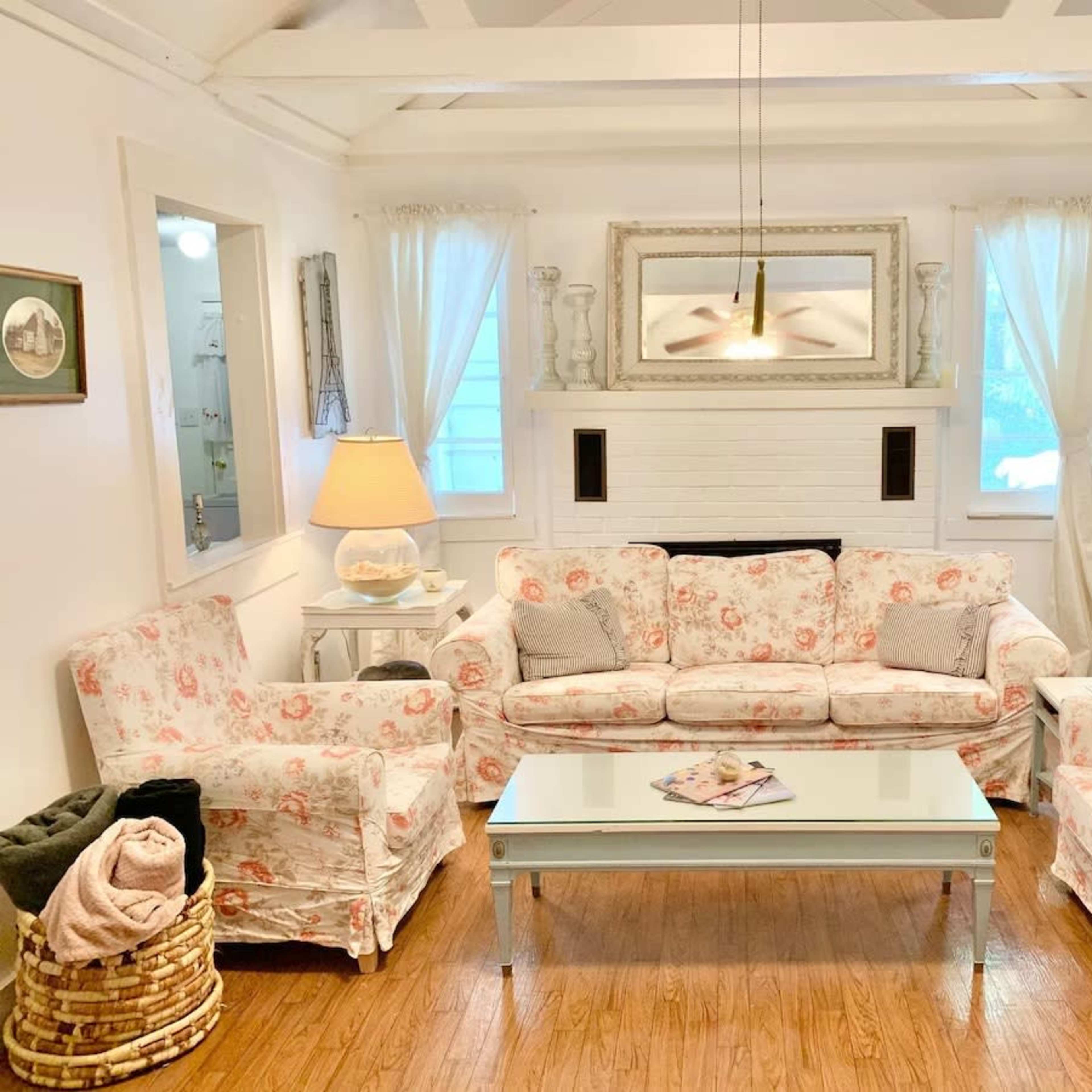 A cozy living room with floral-patterned furniture, a glass coffee table, and large windows allowing natural light.
