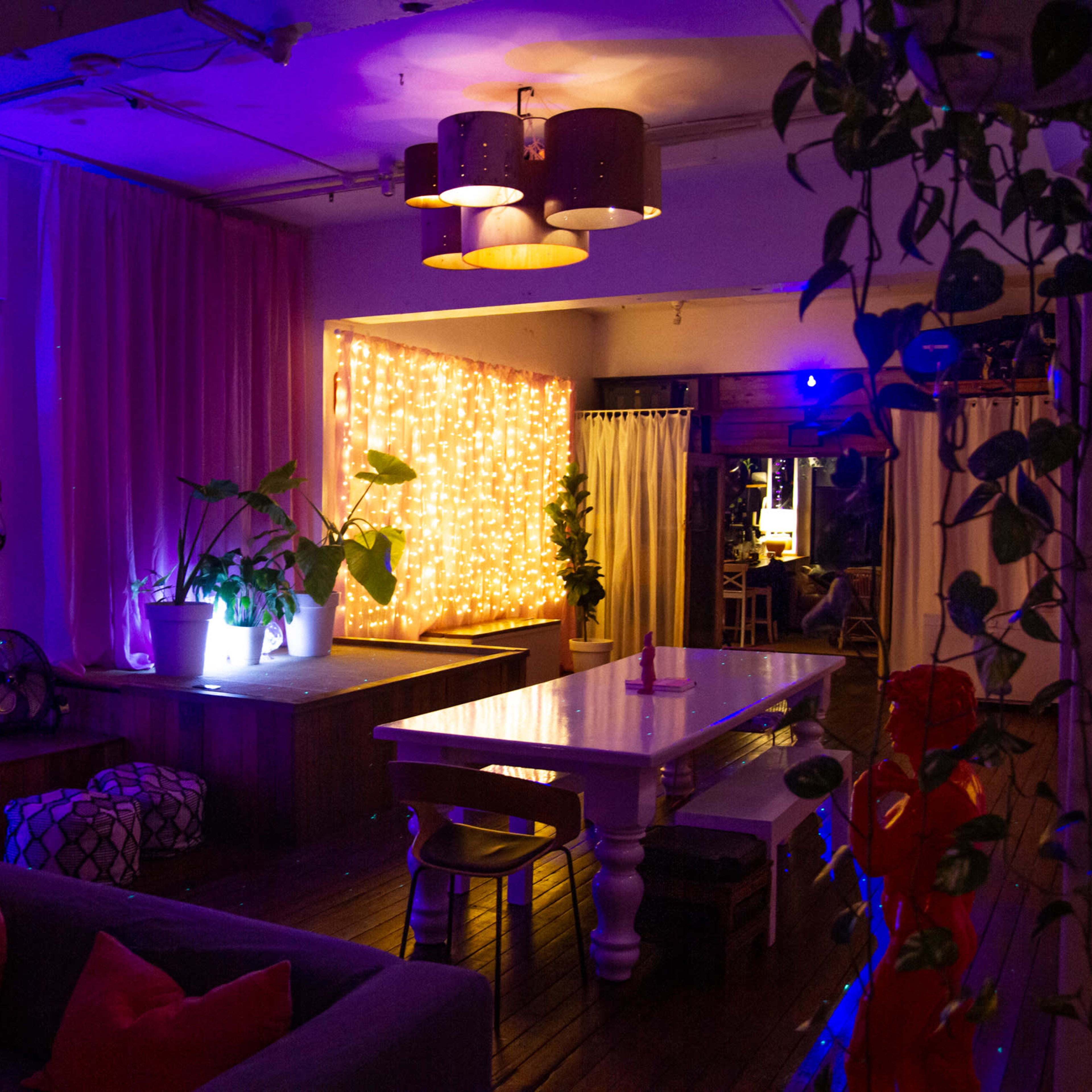 A cozy interior space featuring a large table, soft seating, and decorative lights along the walls.