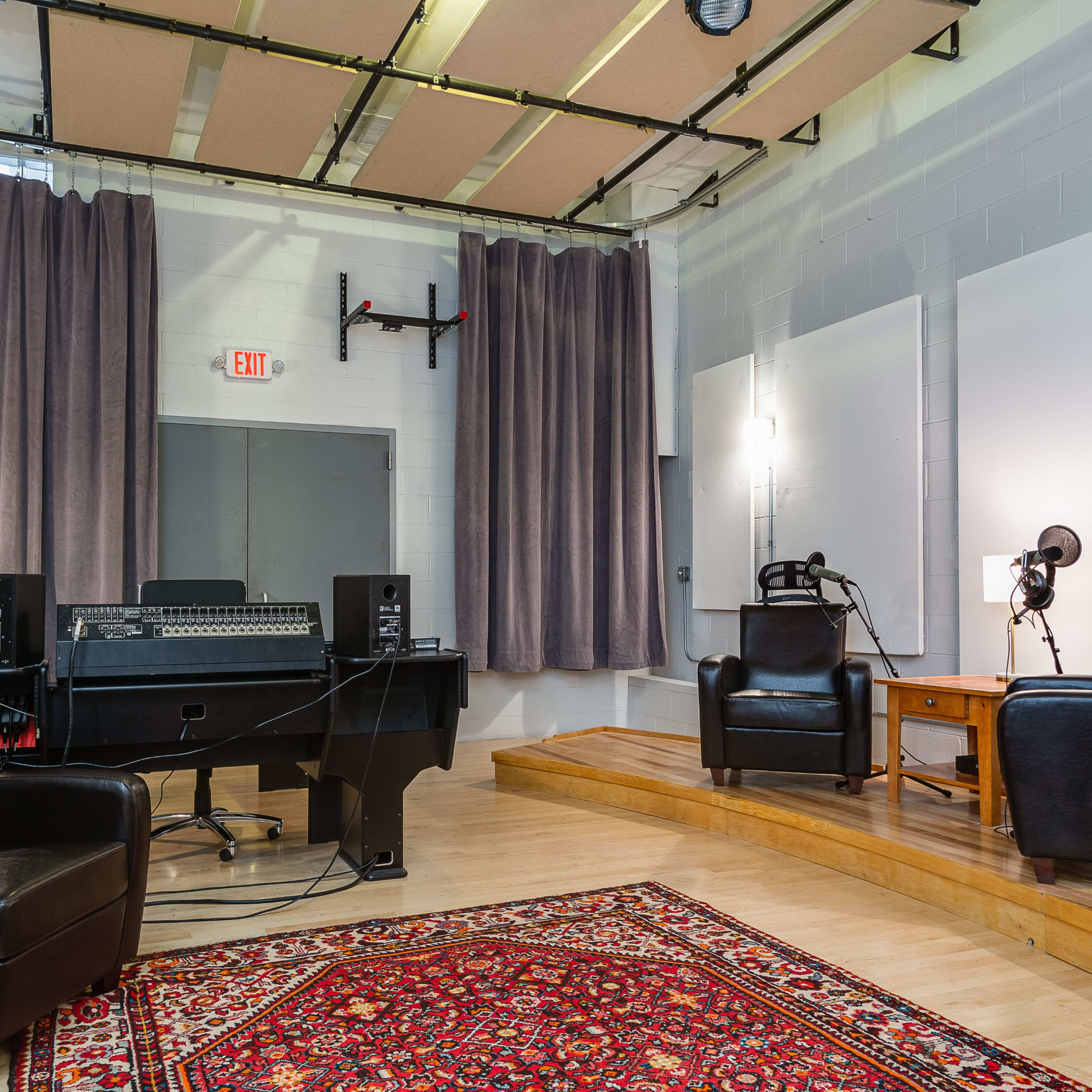 A recording studio with two black leather chairs facing a wooden table and a mixing console, surrounded by soundproof curtains and acoustic panels.