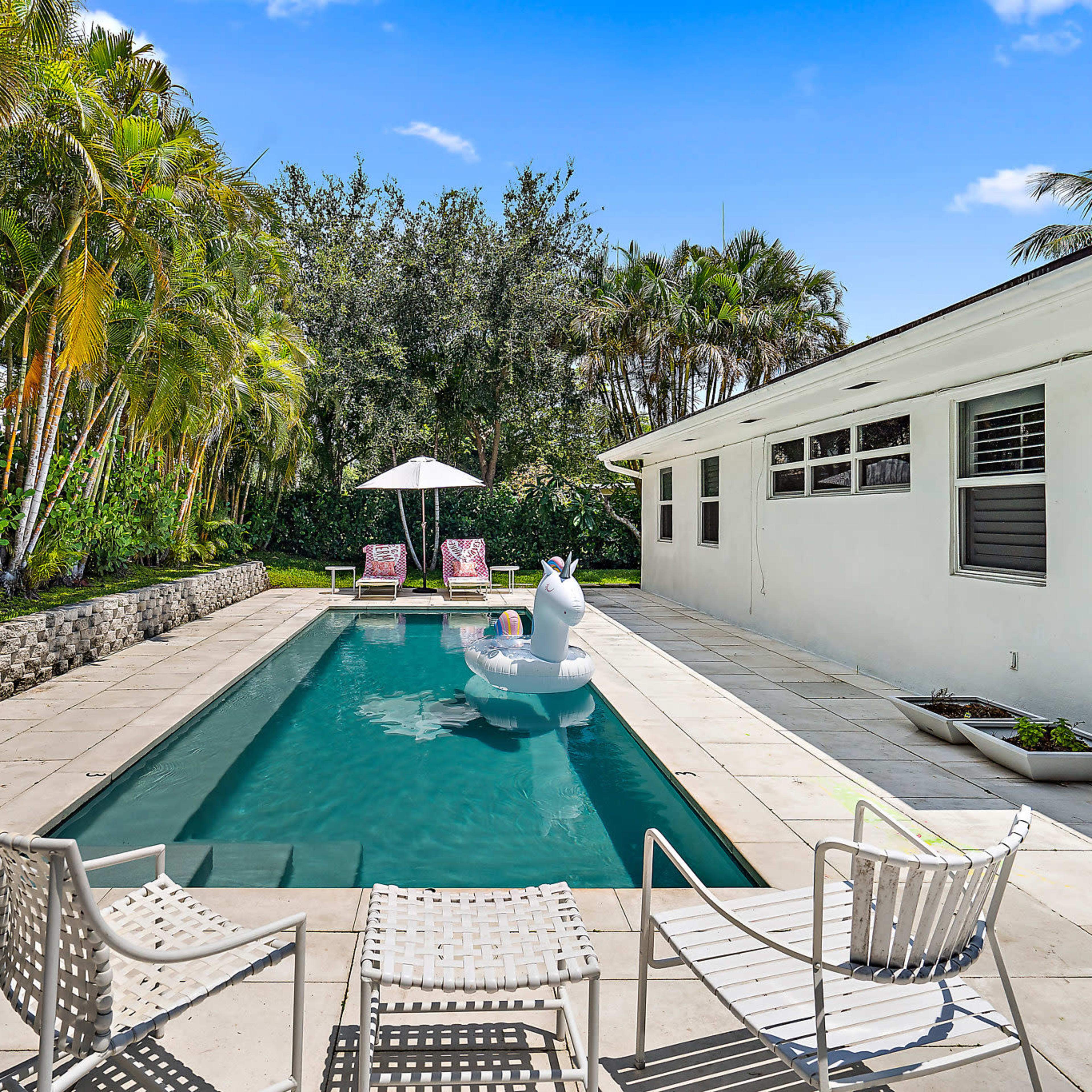 A backyard features a swimming pool surrounded by lounge chairs, palm trees, and a shaded seating area with two chairs and an umbrella.