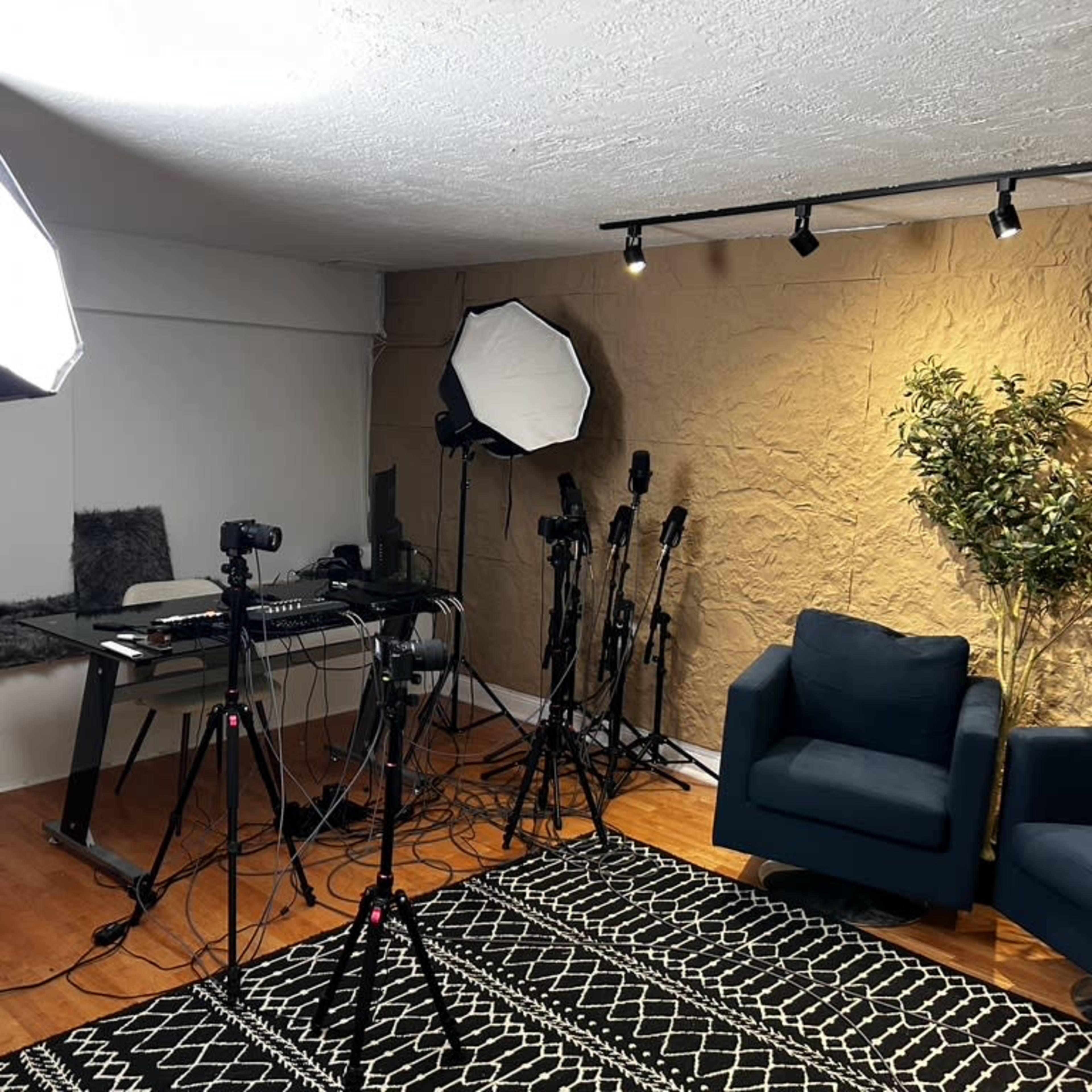 A well-lit studio setup featuring multiple camera tripods, softbox lights, and a seating area with two blue chairs on a patterned rug.