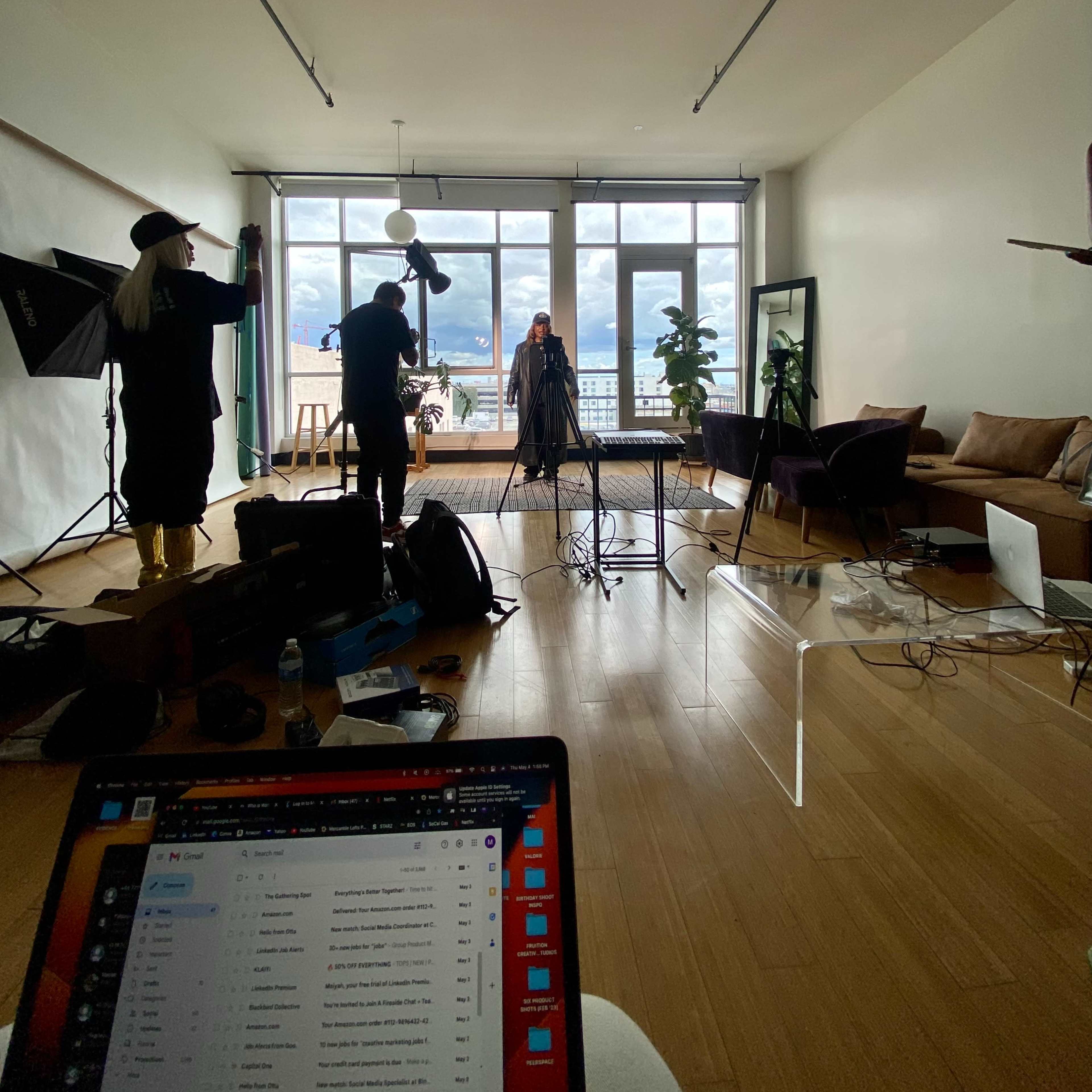 A photoshoot is taking place in a spacious studio apartment with large windows and a view, featuring a subject in front of a backdrop and two photographers with equipment.