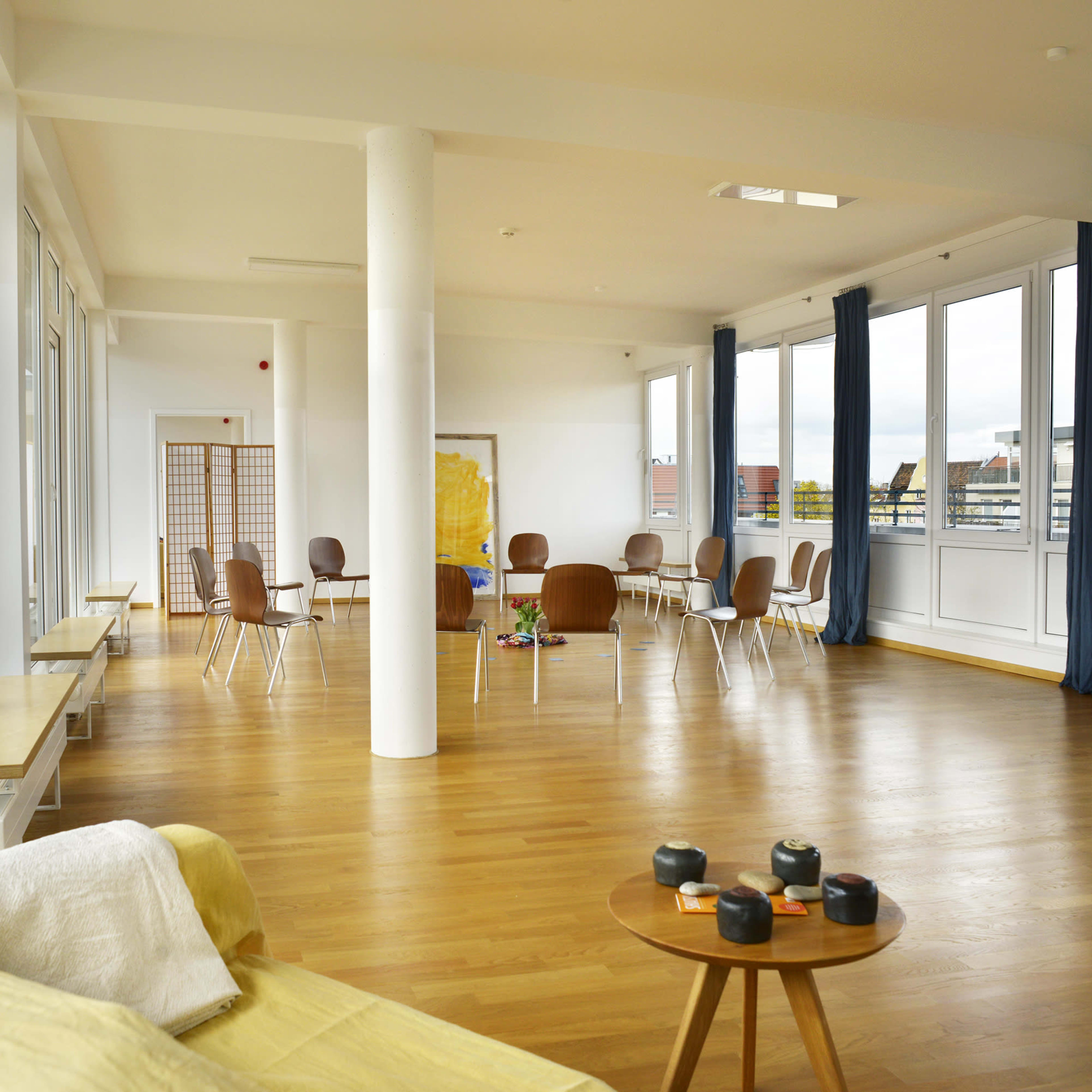 A bright, spacious room with wooden floors, large windows, and arranged seating around a central table.