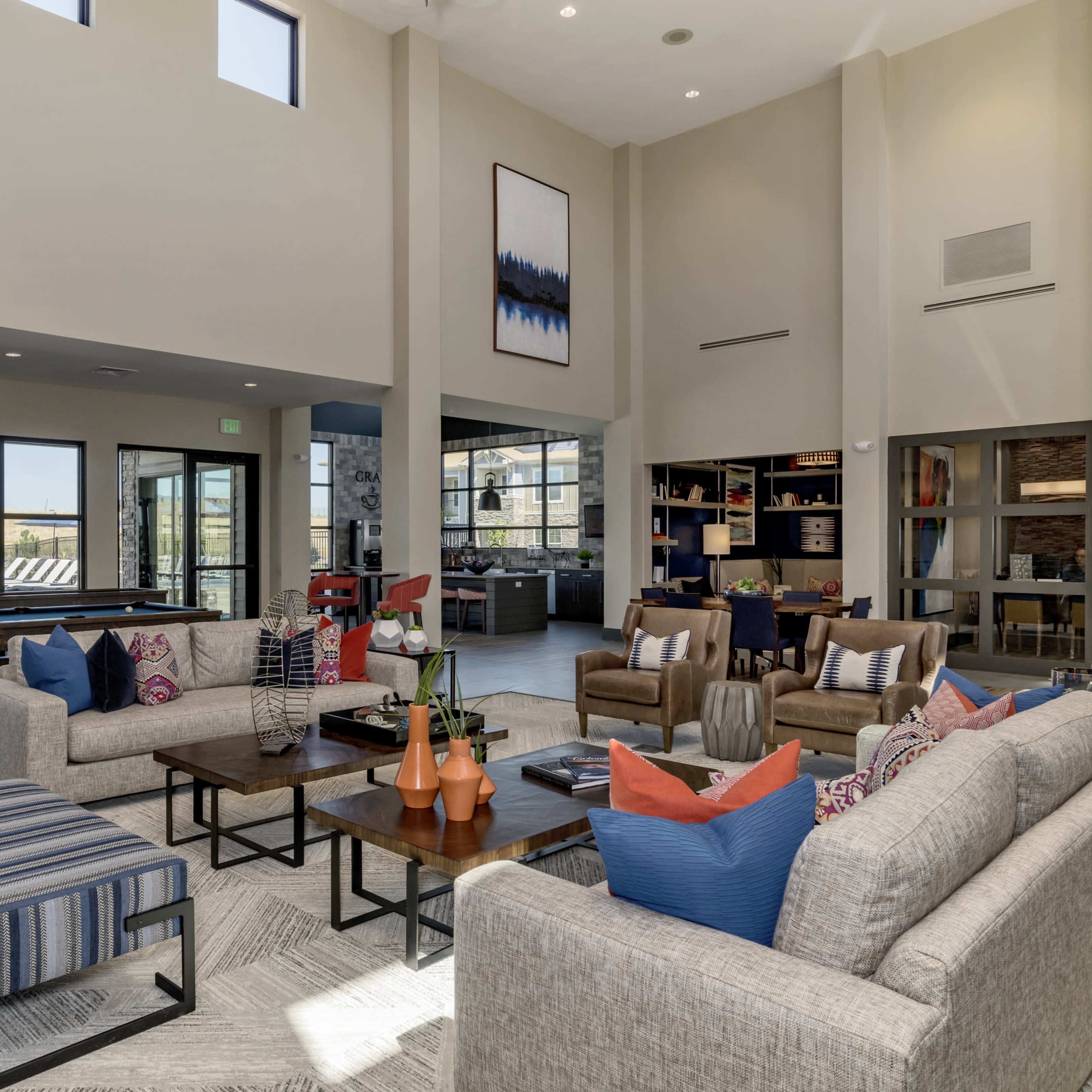 A modern community lounge with a high ceiling, featuring sofas, armchairs, and large windows that provide a view of the outdoors.