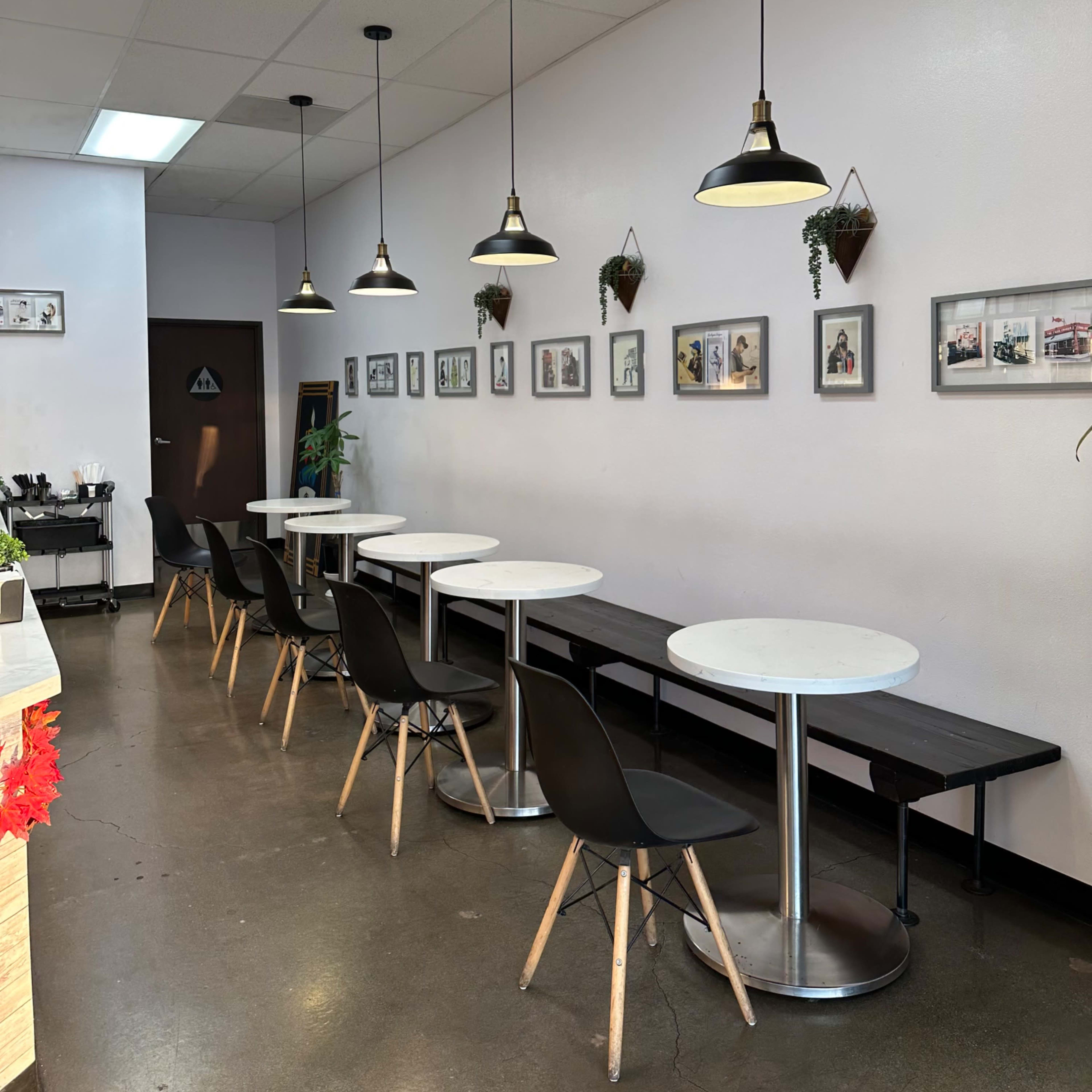 A bright café interior with several white tables, black chairs, and wall-mounted framed photographs.