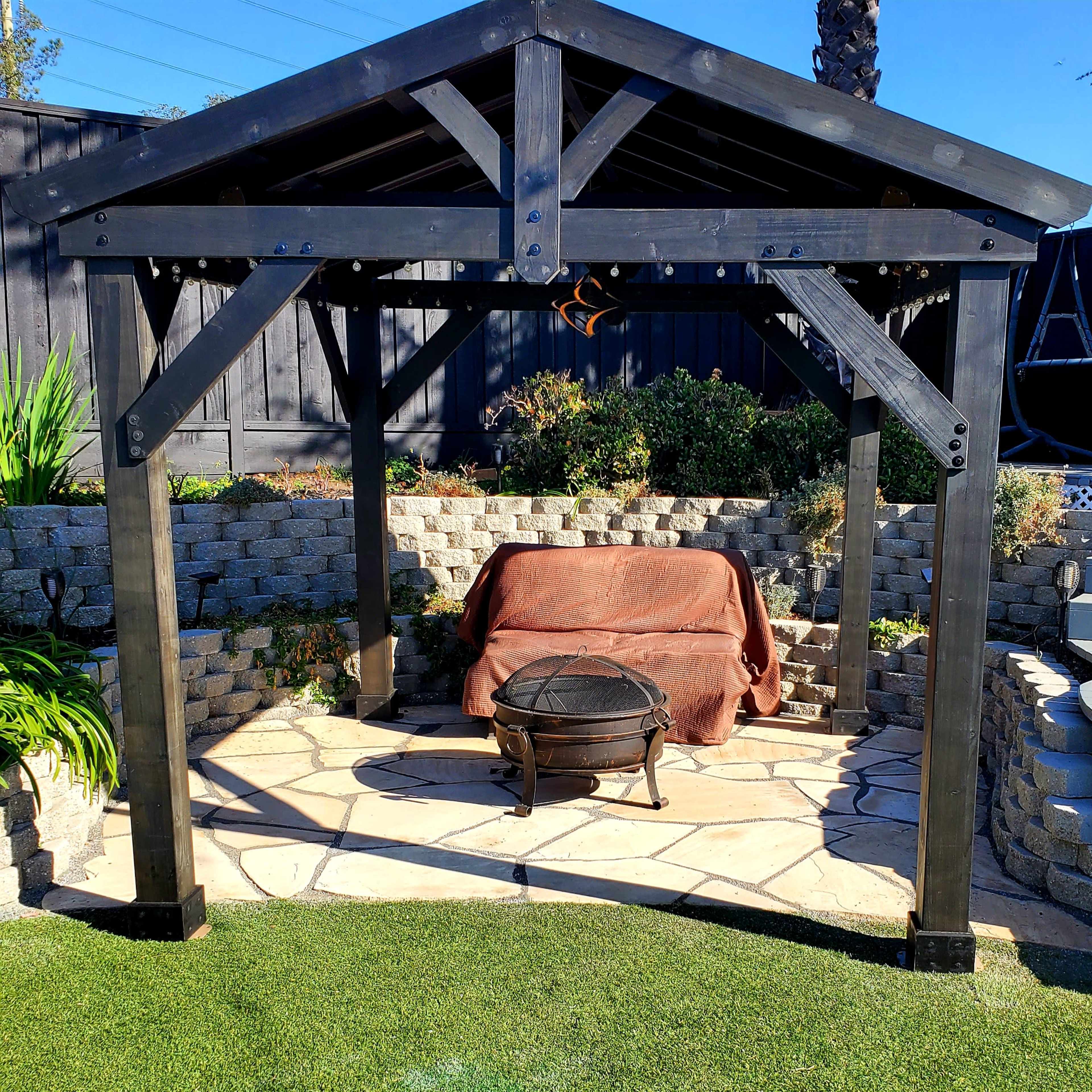 A wooden gazebo with a stone patio and a fire pit stands in a landscaped backyard.