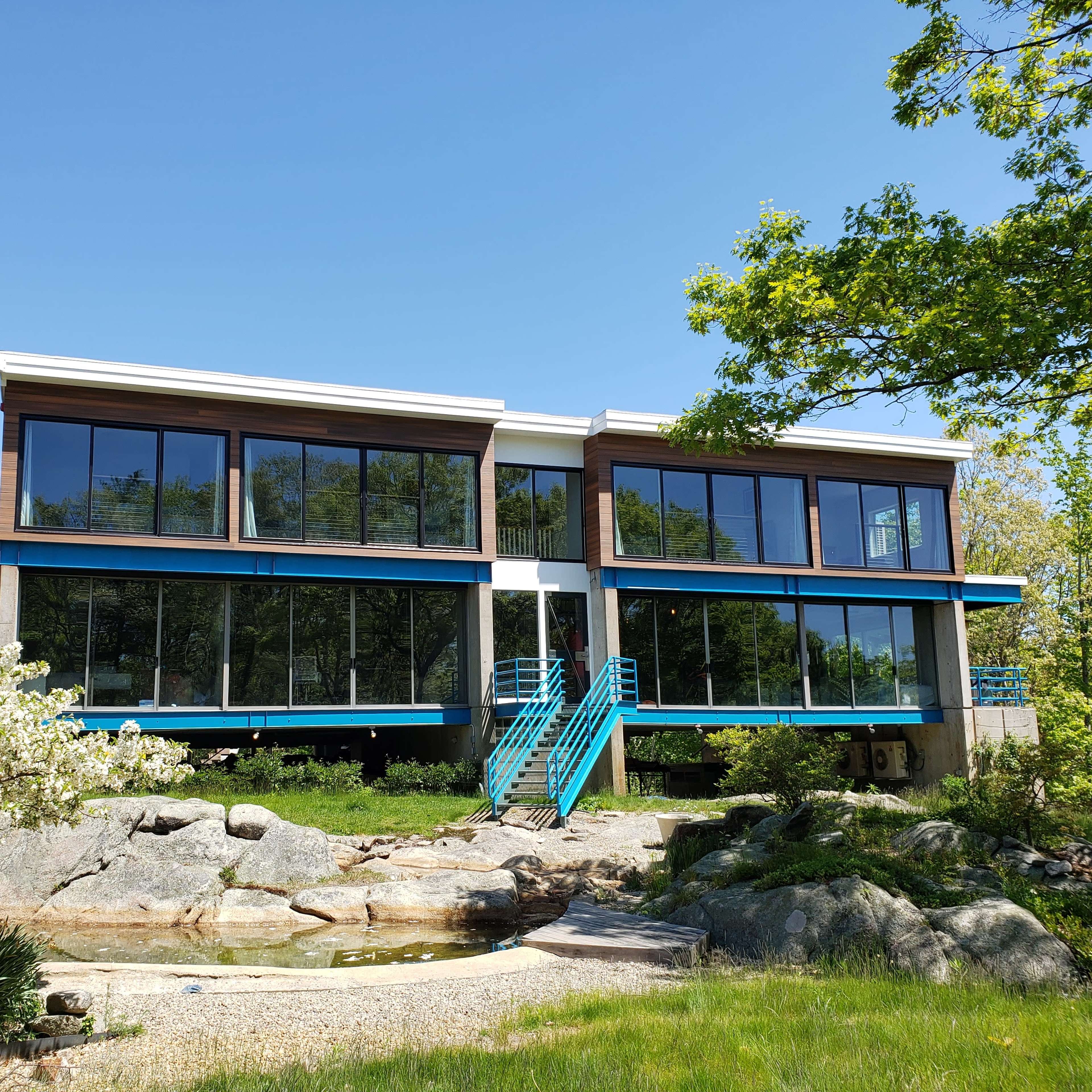 A modern, two-story house with large glass windows and a blue staircase is set amidst greenery and boulders.